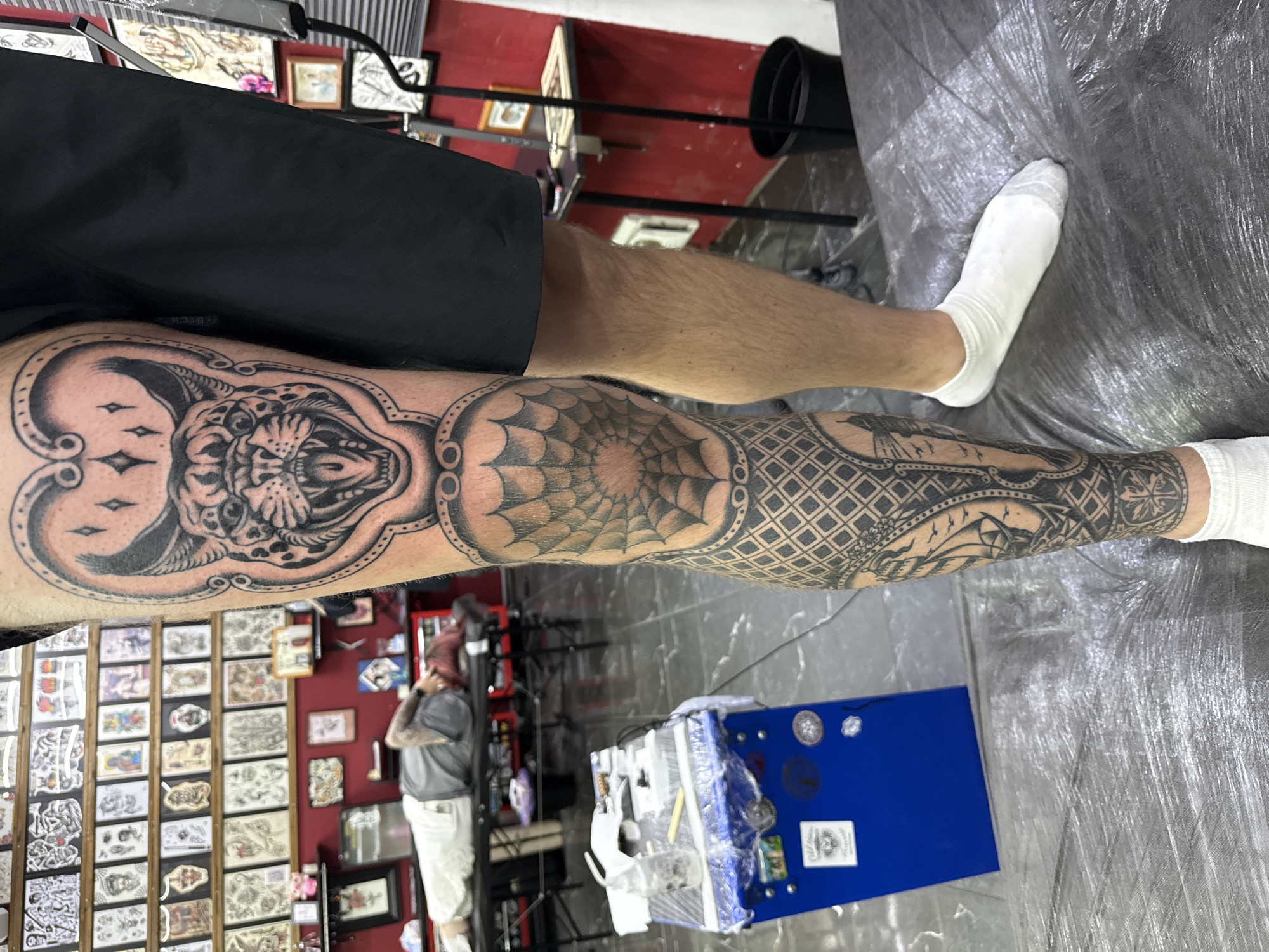 The image captures a close-up view of a person's tattooed leg in what appears to be a tattoo studio. The tattoo features a detailed design of a feline head with its mouth open, set within an ornate frame. Below this, a spiderweb pattern is visible on the knee, transitioning into more intricate geometric and figurative designs further down the leg, including what looks like a ship and nautical elements. The individual is wearing black shorts and white socks. In the background, shelves filled with tattoo flash art line the walls, and a person is lying on a tattoo chair. A blue tattoo machine cart with various supplies is also present. The scene is lit, suggesting an indoor environment. The image does not contain any information that would allow for pinpointing the specific location within Moscow, Russia.