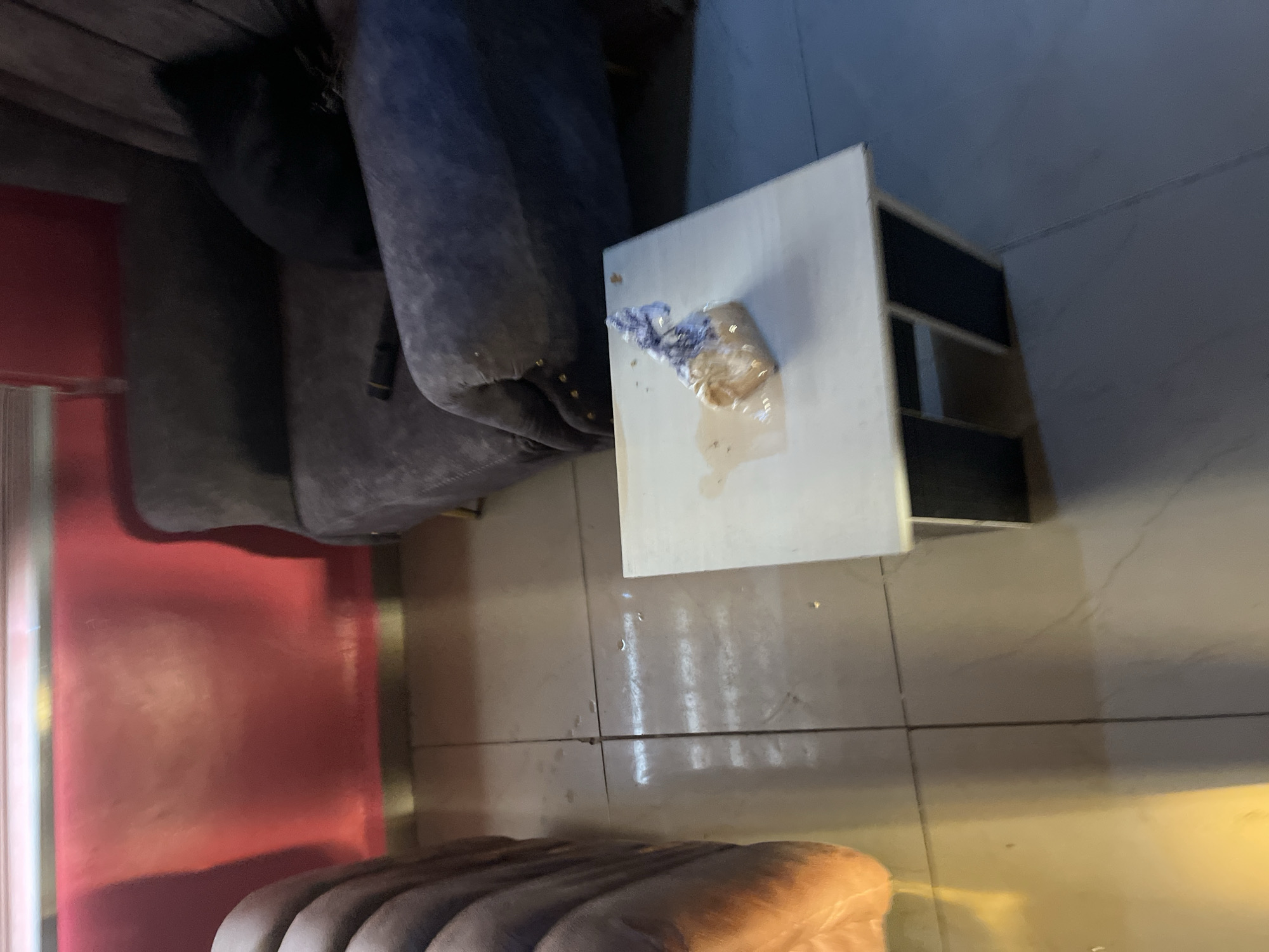 A small, square-shaped coffee table with a light-colored top and dark-colored legs is positioned on a tiled floor. On the table, there is a white plastic bag containing what appears to be food, with a brown liquid spilled around it. The table is situated in an indoor setting, with a plush, dark-colored armchair visible behind it. To the left of the table, a portion of a padded seating area, possibly a sofa or another armchair, is visible. The background features a reddish-pink wall with a subtle metallic sheen. The scene appears to be in a living room or lounge area. The image does not contain any specific indicators that would place it in Jos, Nigeria.