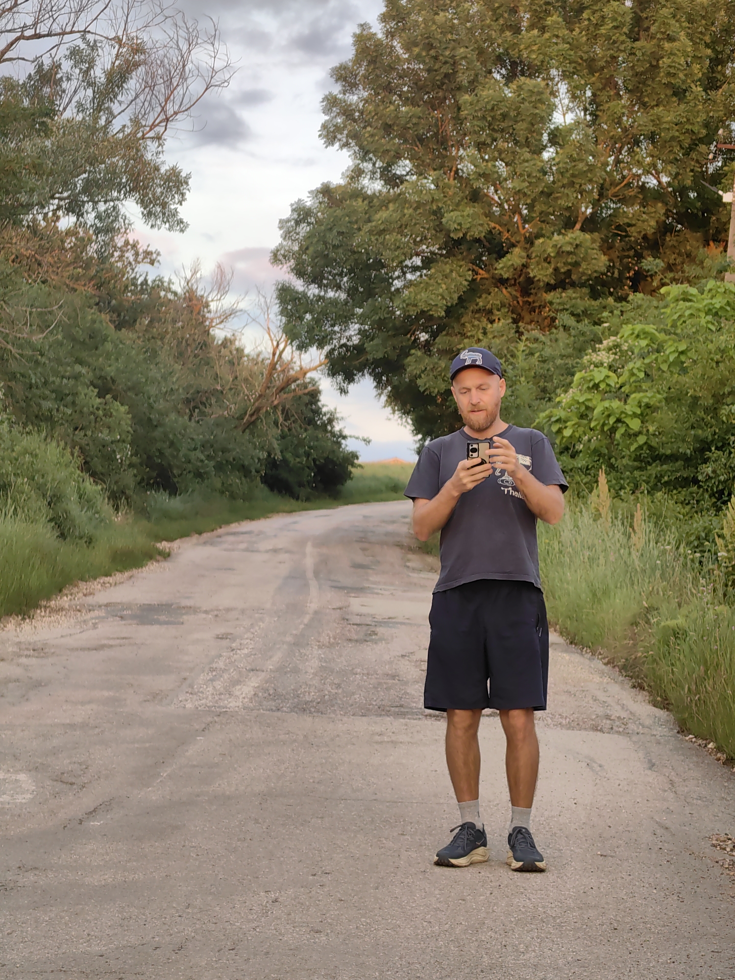 A man stands in the middle of a dirt road in a rural area, looking at his smartphone. He is wearing a dark t-shirt, dark shorts, grey socks, and dark athletic shoes. A baseball cap is on his head. He is surrounded by lush green trees and tall grass on either side of the road. The sky above is partly cloudy with hints of pink and orange from the sunset. The road appears to be unpaved and has some patches of gravel and dirt. The location is described as Ароматновское сельское поселение, Ukraine.