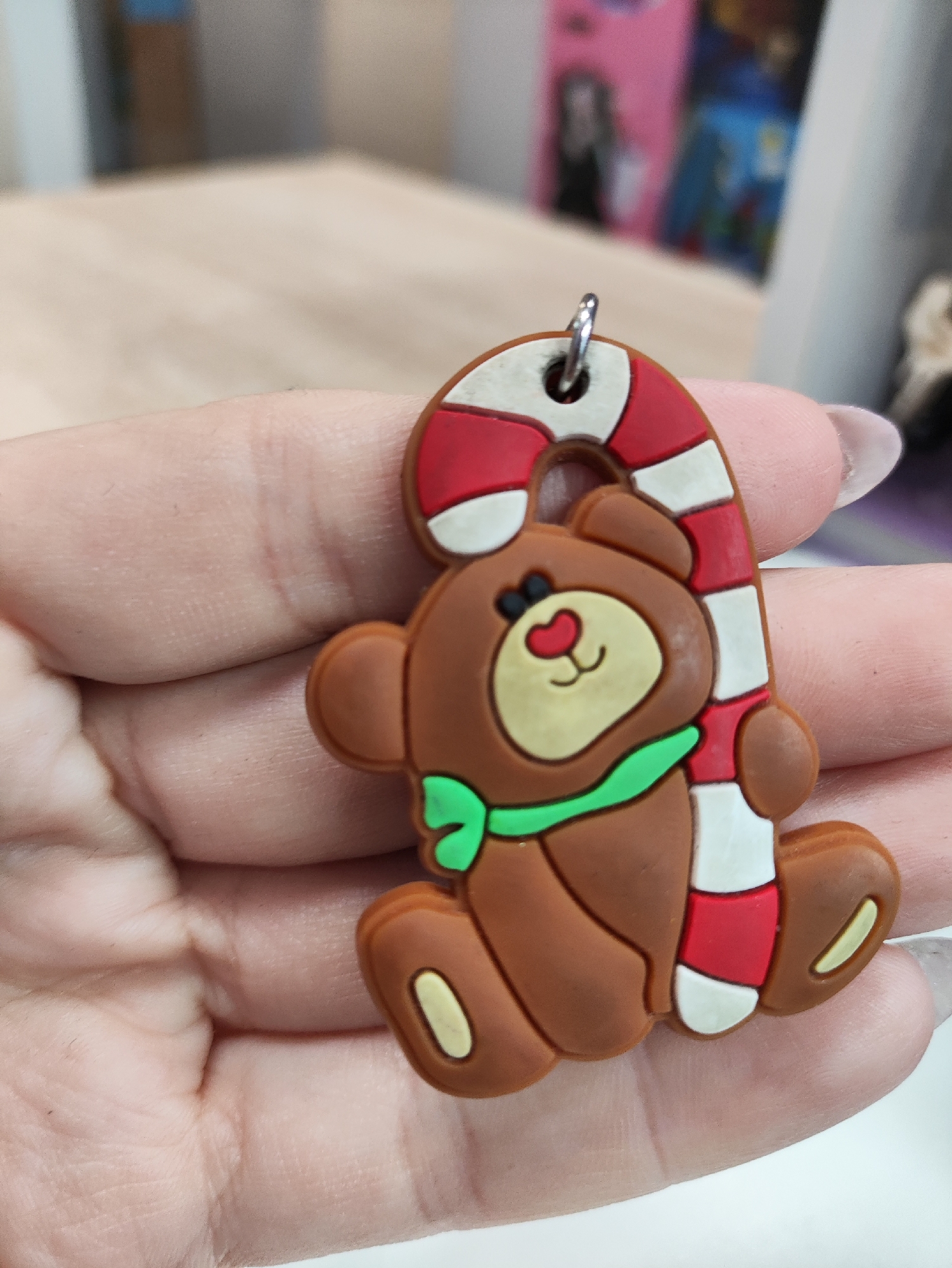 A close-up shot shows a person's hand holding a Christmas-themed keychain. The keychain features a brown teddy bear wearing a green scarf, and it is leaning against a red and white striped candy cane. The background is blurred, showing a wooden surface and some colorful objects, possibly in a store or home environment, but no specific location in the city of Niva, Russia, can be identified from the image.