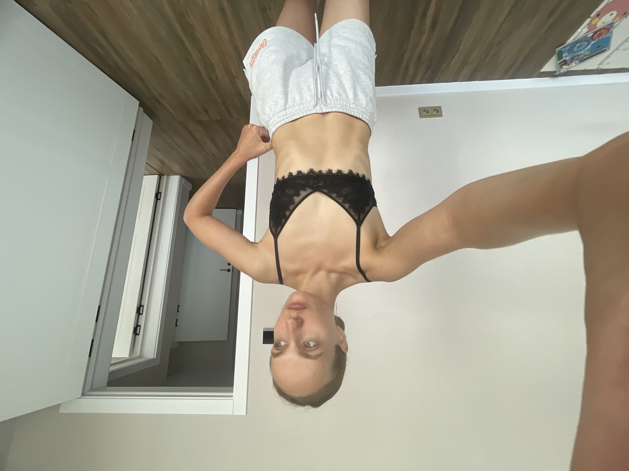 A young woman with blonde hair pulled back stands in what appears to be a residential interior, possibly a bedroom or living area. She is wearing a black lace bralette and light gray sweatshorts with orange lettering. Her left arm is extended towards the camera, obscuring part of her torso, suggesting she is taking a selfie. Her right hand is placed on her hip. The background shows a neutral-colored wall, a wooden floor, and an open doorway leading to another room or hallway with multiple doors visible. Electrical outlets are present on the wall. There is a colorful play mat on the floor in the lower left corner. The lighting suggests it is daytime. The location is identified as Podshivalovo, Russia, though this detail cannot be visually confirmed from the image itself.