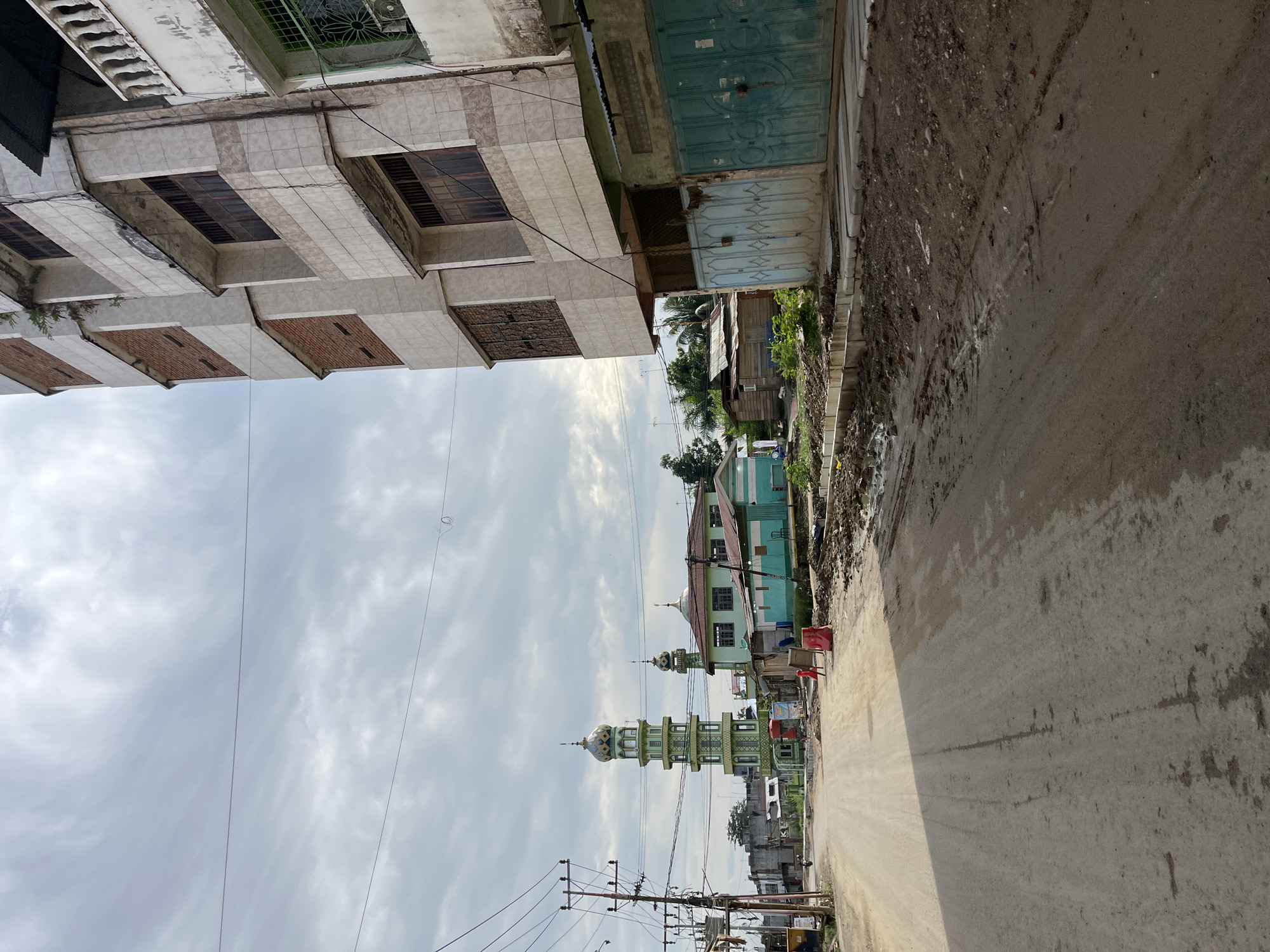 The image depicts a street scene in Kota Kuala Simpang, Indonesia, under an overcast sky. A dirt road, showing signs of recent rain with muddy patches and tire tracks, leads into the distance. On the right side of the road, a multi-story building with white tiled facade and visible balconies is prominent. Adjacent to it, a turquoise building with a metal gate is visible. Across the street, a tall, green minaret of a mosque stands out, accompanied by a smaller mosque building painted in light blue and green. Utility poles with numerous wires are present on the left side of the road, suggesting an urban or semi-urban environment. In the foreground on the right, construction materials or debris are piled up. Further down the road, more buildings and trees can be seen, indicating a developed area. There are a few people visible in the distance, near the mosque, and a red construction barrier on the road. The overall impression is one of an active, developing community with elements of both modern infrastructure and traditional architecture.