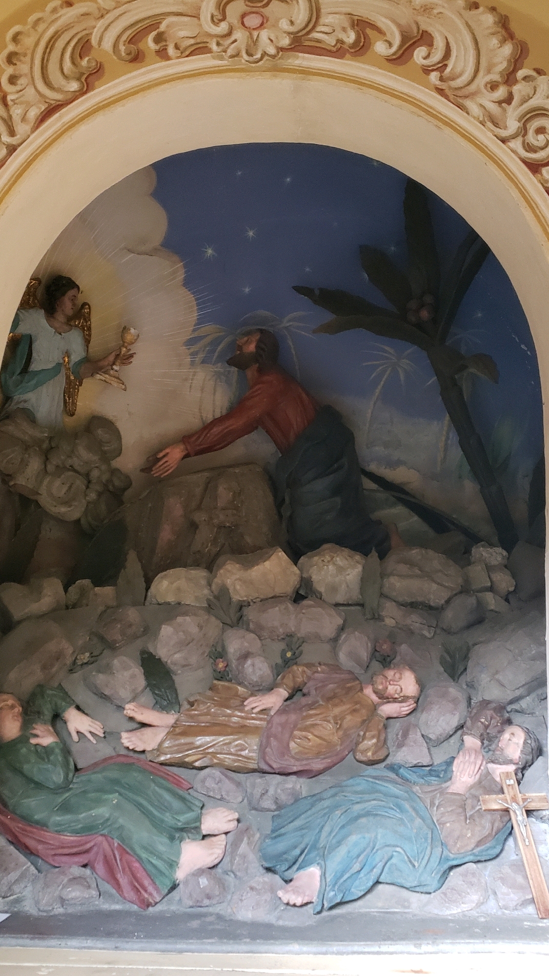 This is a photograph of a relief sculpture depicting a religious scene, likely the Agony in the Garden. The sculpture is located within a niche in a building.
The scene features several figures. In the upper left, an angelic figure with wings and a halo is holding a chalice. To the right, a kneeling male figure in a red garment is reaching out. In the foreground, three figures are depicted lying down amongst rocks. One figure on the far left wears a green garment and is propped up on their left arm. Another figure in a tan garment is lying on their back with their head resting on their arm. A third figure in a blue garment is lying on their back with their hands clasped. A wooden cross is positioned next to this third figure. The background shows a blue sky with stars and palm-like vegetation.
The sculpture is situated within an arched niche that is decorated with ornate stucco work. The overall style suggests a religious art piece. While the precise location in Prešov, Slovakia is not ascertainable from the image alone, it is likely part of a church or a religious building.