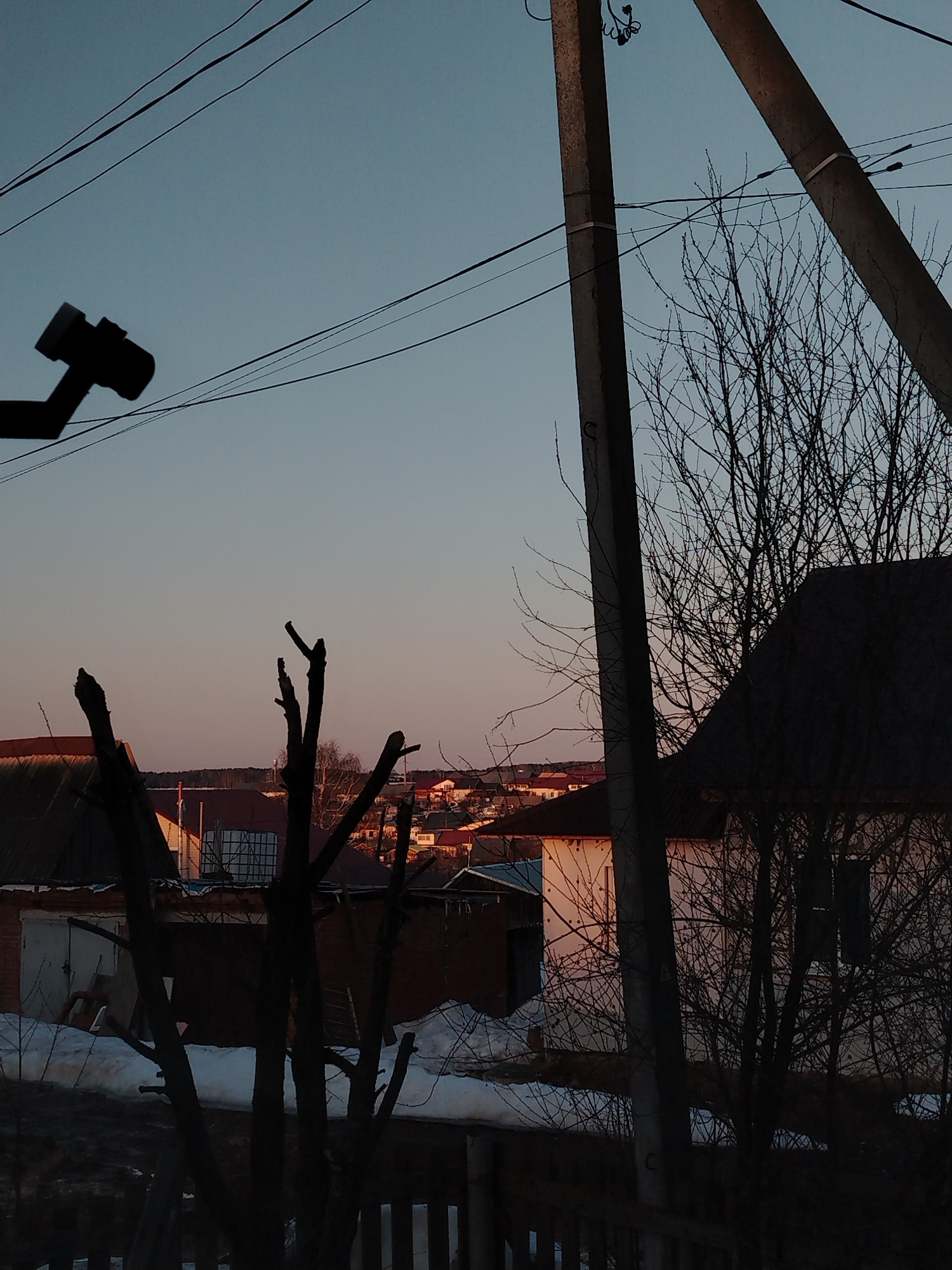 The image depicts a view in Фоки, Russia, capturing a residential scene at dusk. In the foreground, silhouetted bare tree branches and a wooden fence create a foreground frame. Behind these, a snow-covered area is visible next to buildings, including what appears to be a garage and a house. The house has a dark roof and light-colored walls. In the middle ground, a row of other buildings and houses can be seen, receiving the warm light of the setting sun. Dominating the upper portion of the frame are power lines and two utility poles. A security camera is visible on the left side. The sky transitions from a light orange near the horizon to a pale blue further up. The overall impression is of a quiet, possibly rural or suburban neighborhood.