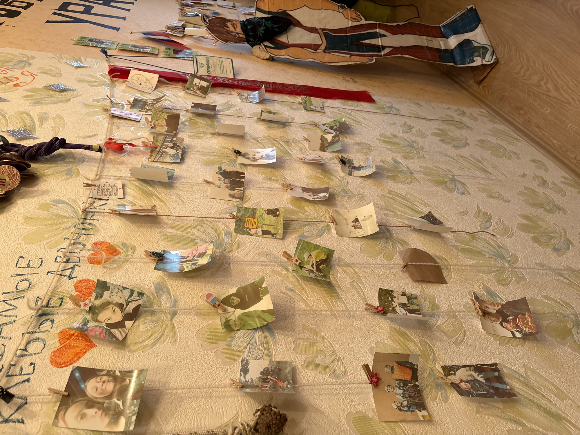 A close-up, slightly angled shot shows a wall in what is likely a room. The wall is covered in wallpaper with a light background and a pattern of stylized leaves. A string art display is stretched across the wall, consisting of multiple horizontal strands of string. Small, folded photographs and paper cutouts are clipped to the string with miniature clothespins. Some of the photos appear to depict people.
At the top of the wall is a cutout of a male character with painted details. Above the string art display is writing in Cyrillic. A red ribbon with writing is also displayed.
A partially visible wooden floor is in the upper right corner.
While I can describe the contents, I cannot confirm the location in Chaikovsky, Russia, without additional contextual information.