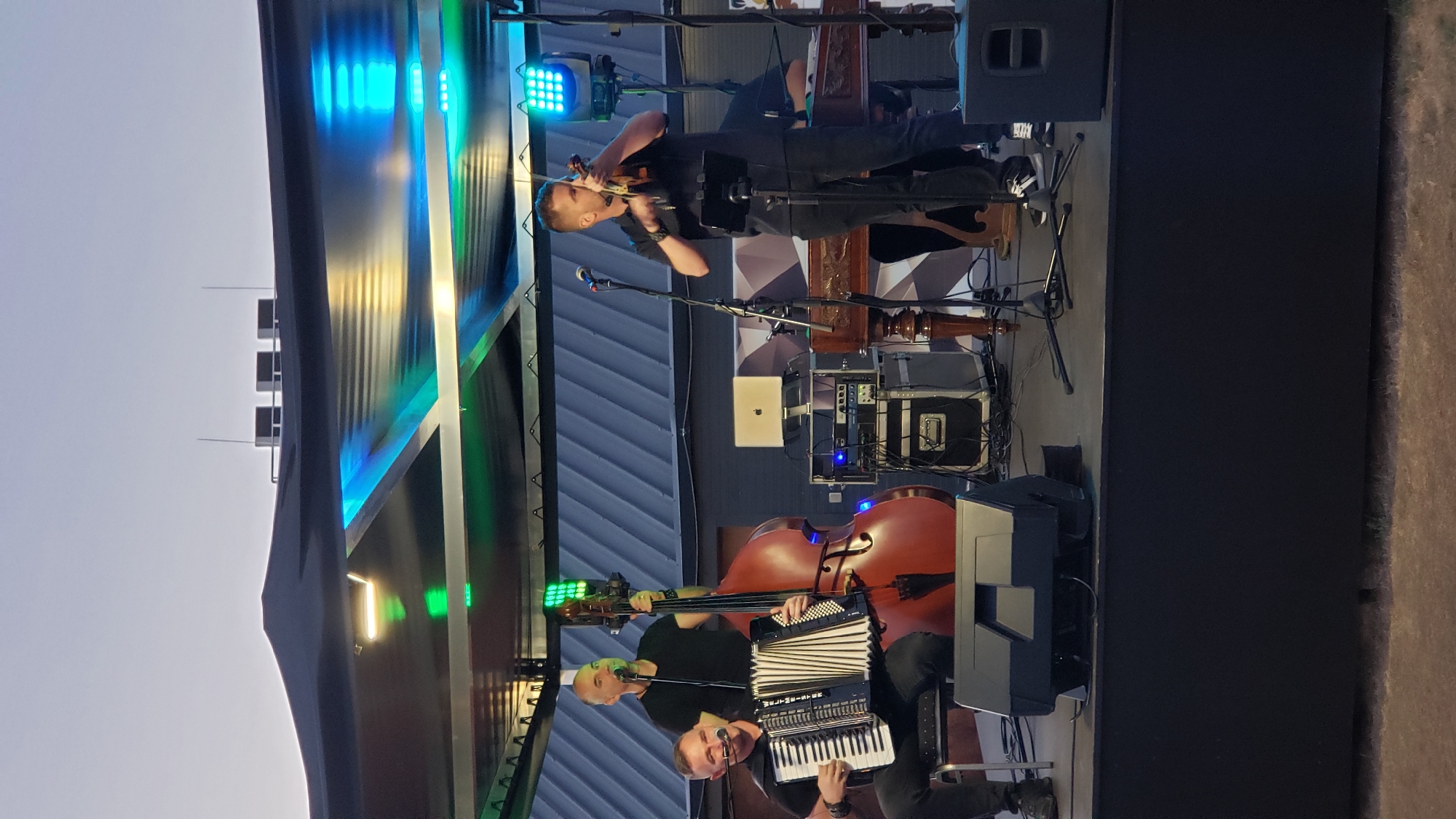 A musical ensemble performs on a stage under a canopy at dusk. The stage is set up outdoors, likely in the city of Prešov, Slovakia, given the context. Three male musicians are visible. On the left, a man sits playing an accordion, wearing a black t-shirt and black pants, with black shoes. Next to him, a man stands playing a double bass, also dressed in black. To the right, another man stands playing a violin, wearing a black shirt and black pants, with black and white sneakers. Behind the violin player, a dulcimer-like instrument is partially visible. The stage is equipped with microphones, audio equipment cases, and a laptop. Colored LED lights are mounted on the canopy, casting blue and green glows. The background shows a muted, grey sky, suggesting either a twilight or overcast condition.