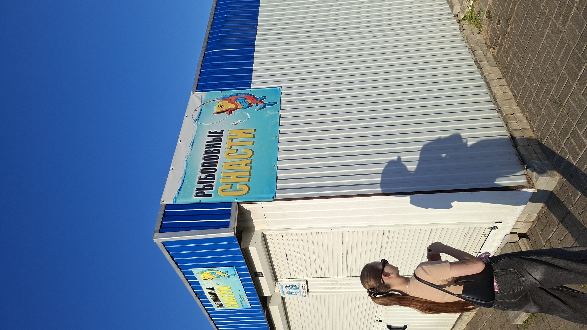 A young woman with long brown hair pulled into a ponytail and wearing sunglasses, a tan t-shirt, and dark wide-leg jeans stands in front of a white, corrugated metal building with a blue trim. She carries a black shoulder bag. A shadow of the woman is cast on the building's wall. A sign on the building reads "РЫБОЛОВНЫЕ СЧАСТИ" in Russian, which translates to "FISHING HAPPINESS". Another smaller sign is visible on the left side of the building. The scene appears to be outdoors, with a clear blue sky above and a paved walkway in the foreground. The building is likely a shop or kiosk. The context suggests this location is in the city of Myadzyel, Belarus, possibly near a body of water given the fishing-themed signage.