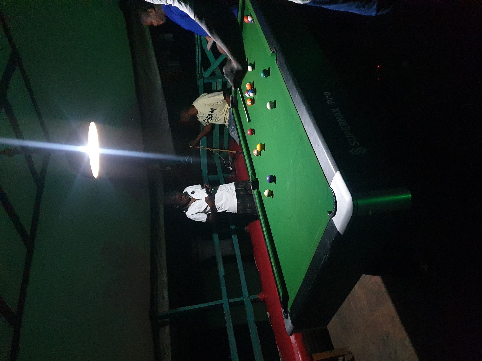 In Lokoja, Nigeria, an outdoor covered area is depicted at night. A group of individuals are engaged in a game of pool. A young person in a white polo shirt stands near the pool table, looking at their phone. Another person, wearing a yellow t-shirt with the word "DREAM" on it, is seated and holding a pool cue. Two other individuals are partially visible on the right side of the frame, one in a blue shirt and another in a white shirt. The pool table is green with several balls on it, including a cue ball. The scene is illuminated by a bright overhead light. The environment includes a dark background, a railing, and a red bench. The pool table has "SUPERMAX Pro" branding on its side.