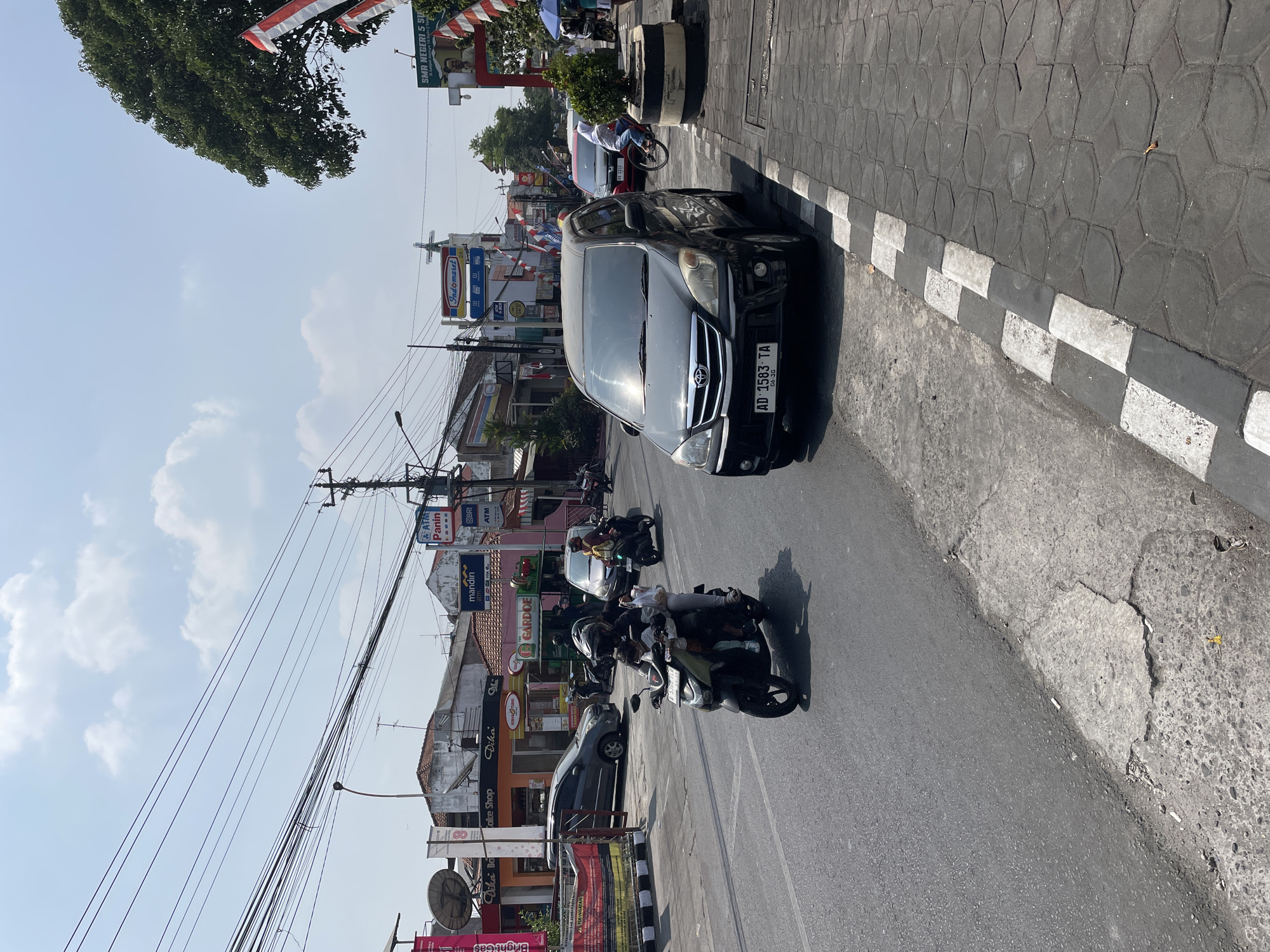 The image depicts a street scene in a city. A black Toyota Avanza is parked on the side of the road. A dark-colored motorcycle with two occupants, a driver and a passenger, is in the foreground, traveling on the road. Several other motorcycles and a car are visible in the background. The buildings lining the street appear to be shops and commercial establishments. Overhead, numerous electrical wires crisscross the sky. The sky is bright with scattered clouds, indicating daytime. The street is paved, and there is a sidewalk on the right side of the image. A large tree with lush green foliage is also visible on the right. Indonesian flags are displayed on poles along the street.