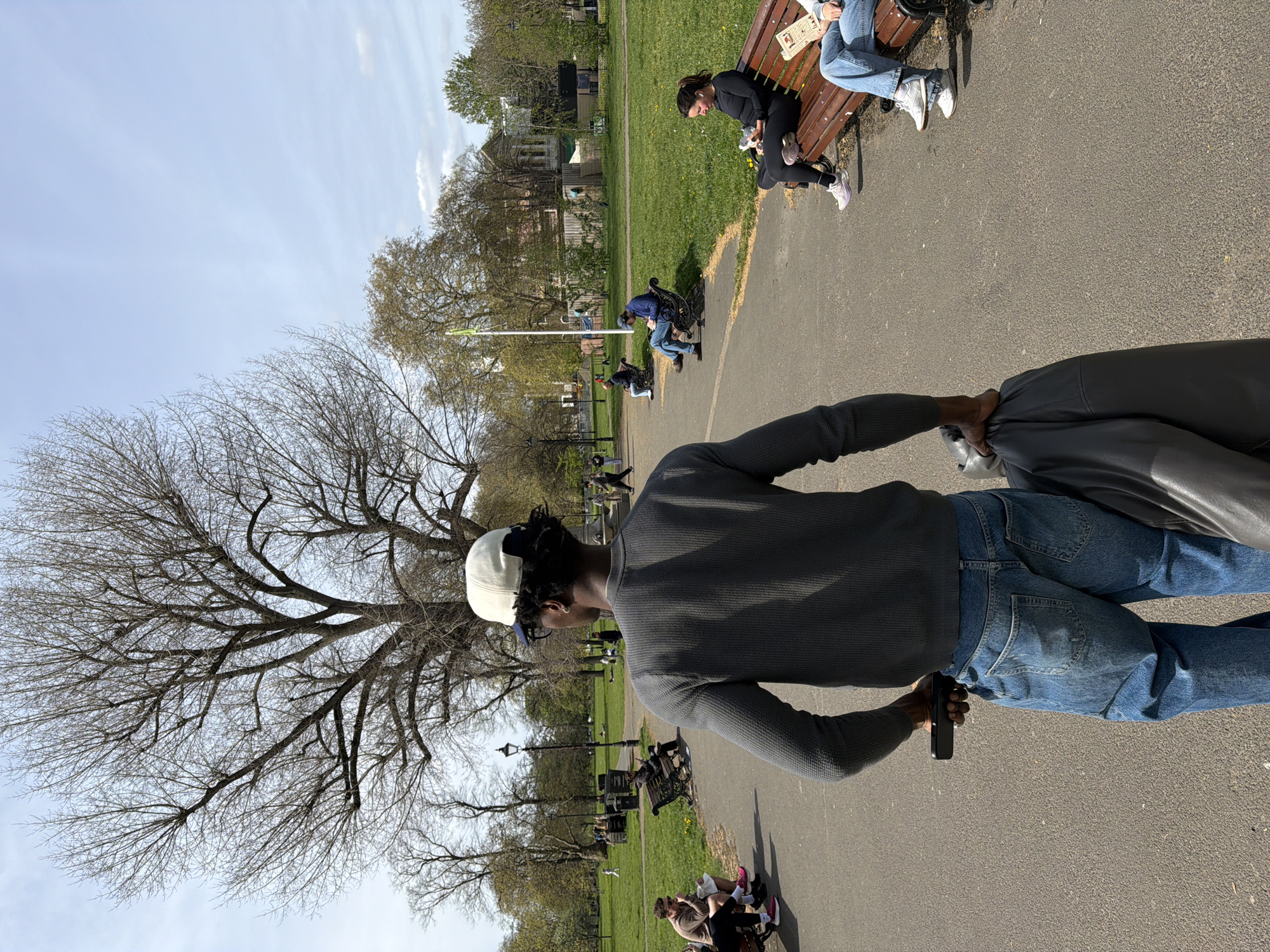 The image captures a scene in a park setting, likely in London, United Kingdom, based on the general architectural style and the park's features. A man is standing on a paved pathway, wearing a grey sweater, blue jeans, and a cap. He is holding a dark-colored device, possibly a mobile phone, in one hand and a grey jacket in the other.
In the background, other people are seated on benches, some engaging in activities like reading or relaxing. The park features trees with bare branches, suggesting it might be early spring or late autumn. Grass covers the ground surrounding the pathway. A building can be seen in the distant background. The sky is blue with scattered clouds.