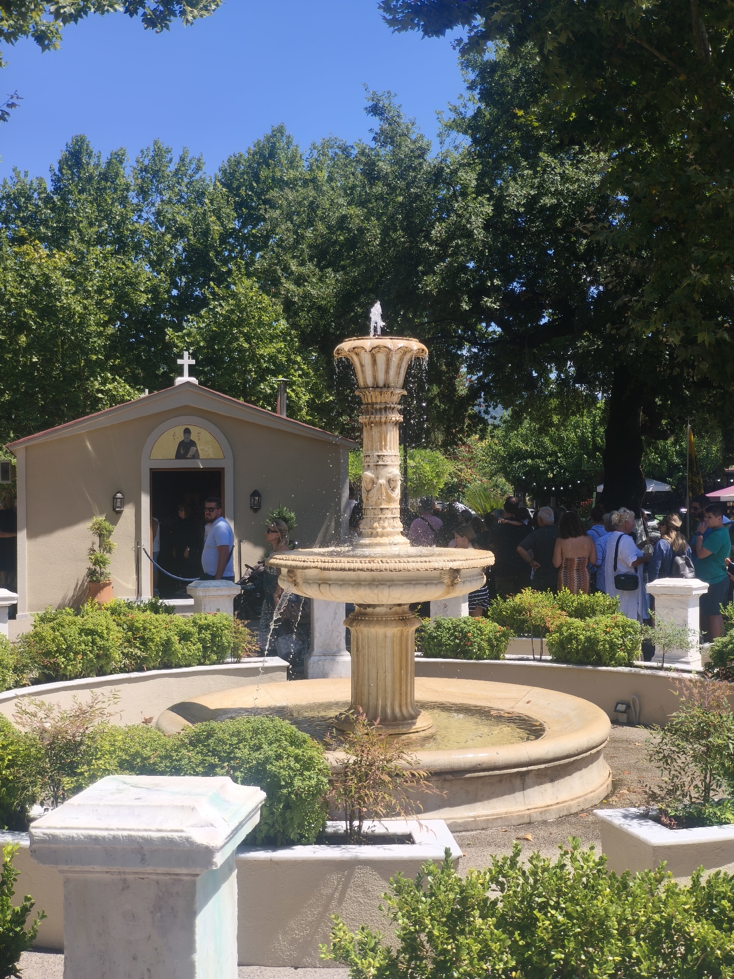 The image shows a courtyard in Prokopi, Greece, with a small chapel and a multi-tiered fountain as the central elements. The chapel, a light beige building with a red-tiled roof, features an arched entrance with a religious icon above it and a cross on its peak. Several people are visible near the chapel entrance, with one man in a blue shirt standing prominently. The fountain, made of a light-colored stone, has water cascading from its upper tier into a circular basin surrounded by lush green bushes. In the background, large trees provide shade, and a crowd of people are gathered, suggesting an event or a popular gathering spot. The overall environment is sunny and outdoors, with a clear blue sky.
