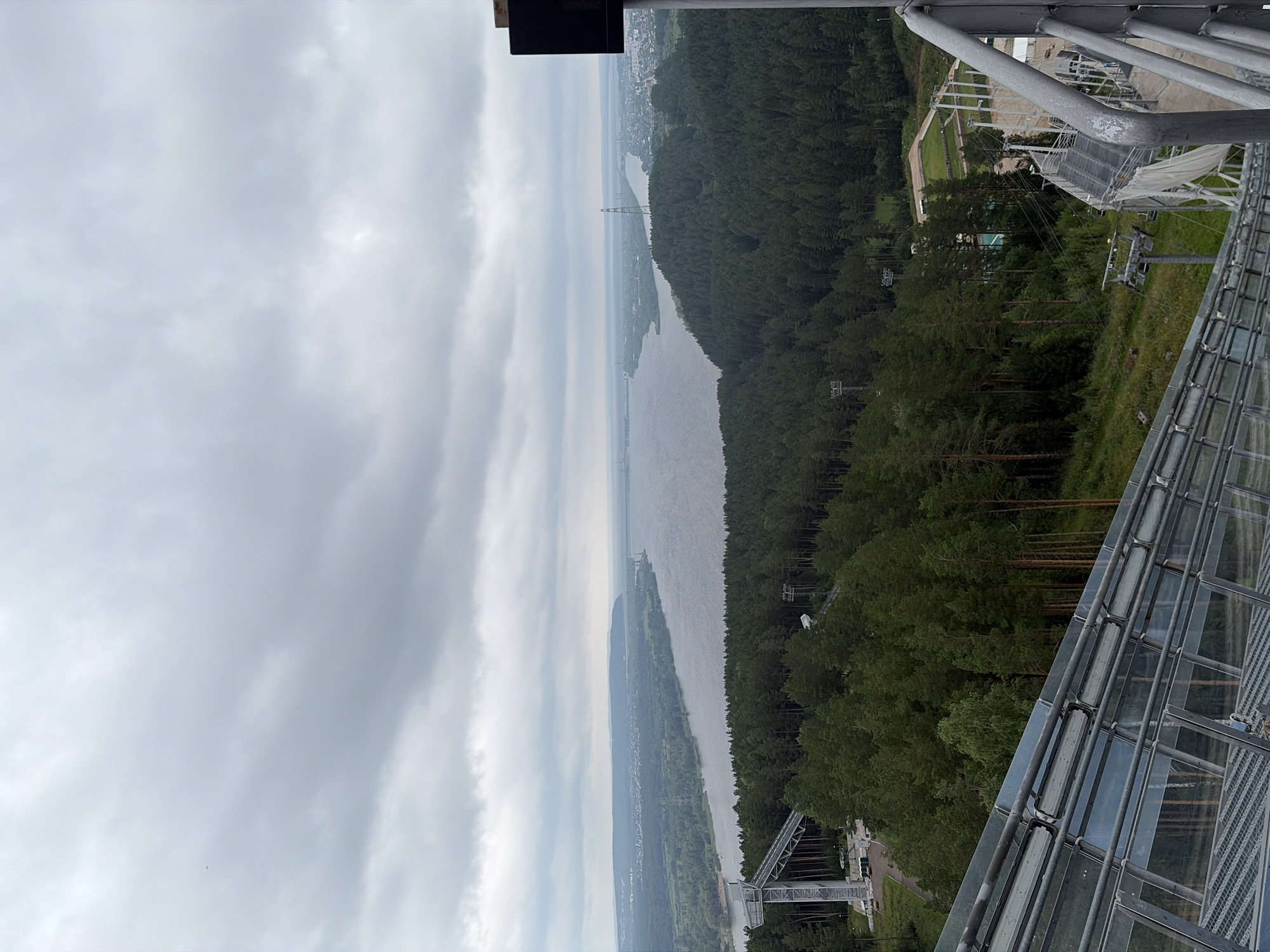 An aerial shot shows a wide, calm river flowing through a landscape of lush green forests and distant urban development under a heavily overcast sky. In the foreground, a modern ski jump structure with a glass-covered track is visible, descending from the viewer's perspective. The ski jump structure is made of metal and glass, with lights mounted along the track. A ski lift system with poles and chairs is also present, operating on the hillside adjacent to the ski jump. Further into the landscape, the river bends, with a bridge crossing it in the distance. Trees dominate the middle ground, creating a dense forest that stretches to the riverbanks. On the far bank of the river, a city or town is visible, with buildings and infrastructure extending towards the horizon. The sky is filled with grey, diffused clouds, suggesting a cloudy or rainy day. A few small, dark figures are visible on the ski jump structure, possibly spectators or workers. The scene appears to be a recreational area or a viewpoint overlooking a natural and urban environment.