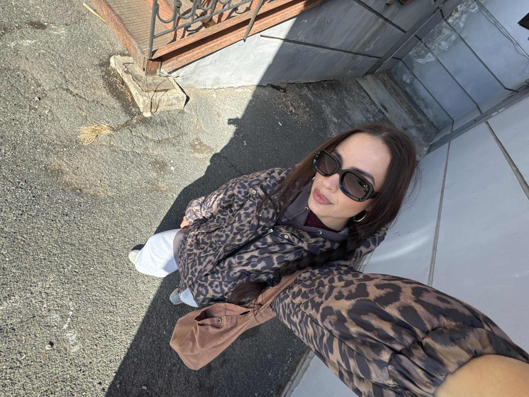 A top-down perspective captures a woman in an urban setting. She dons a leopard-print jacket, sunglasses, and has long brown hair. The ground is paved, with shadows suggesting a bright day. The setting appears to be in Krasnoyarsk, Russia, indicated by the architectural style and urban layout.