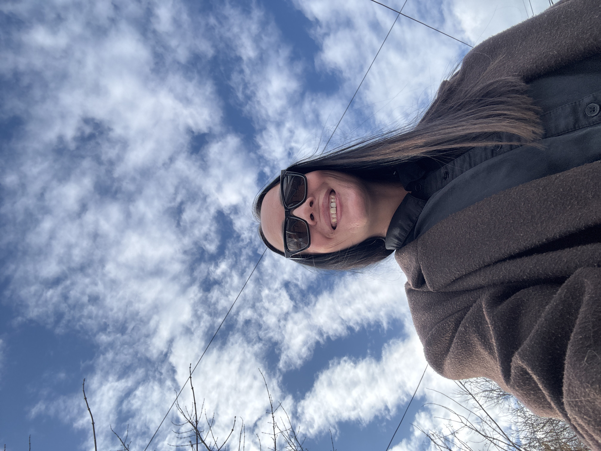 An outdoor, low-angle shot depicts a smiling woman in Chaikovsky, Russia. The subject is positioned in the lower half of the frame, looking directly upwards towards the camera. She wears dark, rectangular-framed sunglasses, a dark button-up shirt, and a textured, dark brown overcoat. Her long, dark hair falls over her shoulders. The background consists primarily of a bright blue sky partially obscured by numerous scattered white clouds. Bare tree branches are visible along the left edge of the frame, and multiple dark utility wires horizontally traverse the upper sky.