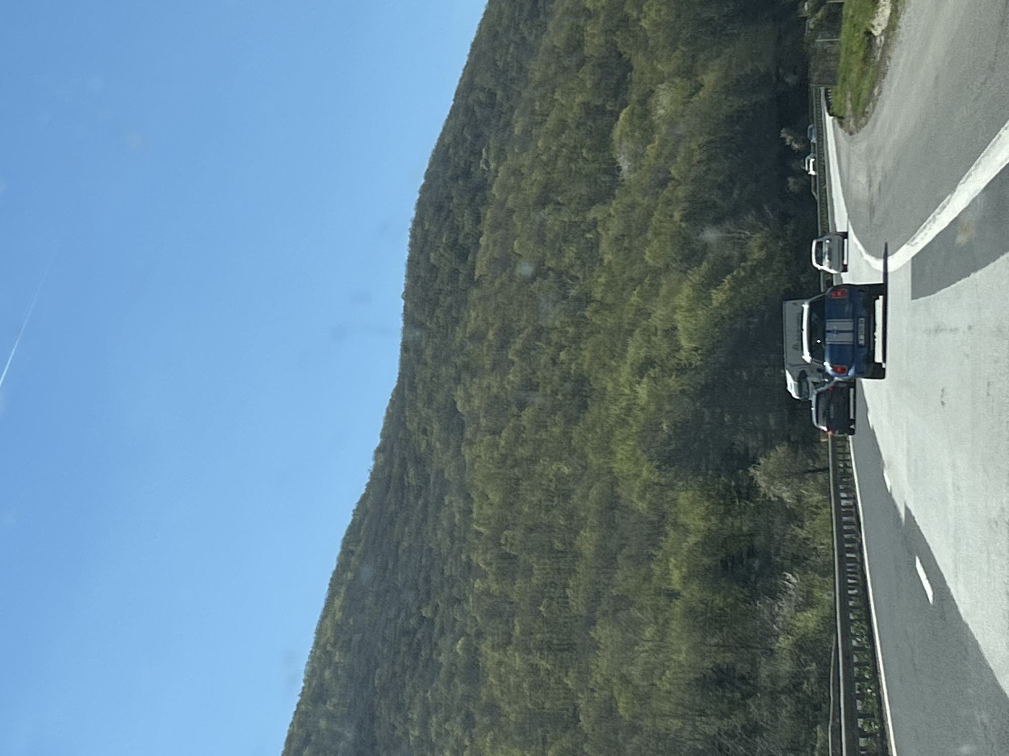The image shows a mountain road near Dolno Varshilo, Bulgaria. The sky is clear and blue. A mountain covered in dense green vegetation runs along the left side of the road. The road has a white line marking the lane division. Several cars are visible on the road. A guardrail separates the road from the mountainside.
