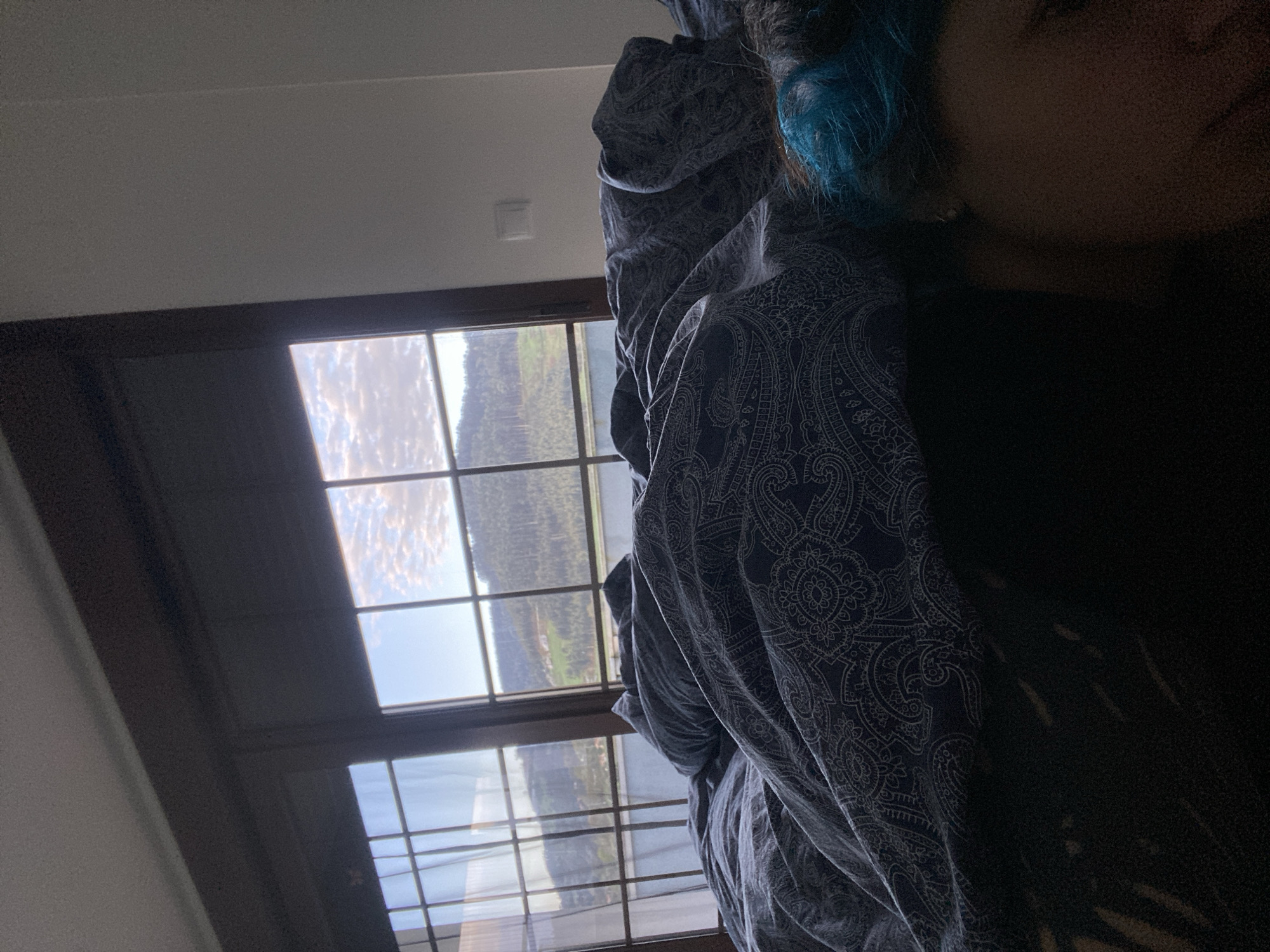 A person with dyed blue hair is lying in bed under a patterned duvet, partially obscured by the duvet. A large window with multiple panes shows a landscape with trees and a cloudy sky. The location is Villaviciosa, Spain.