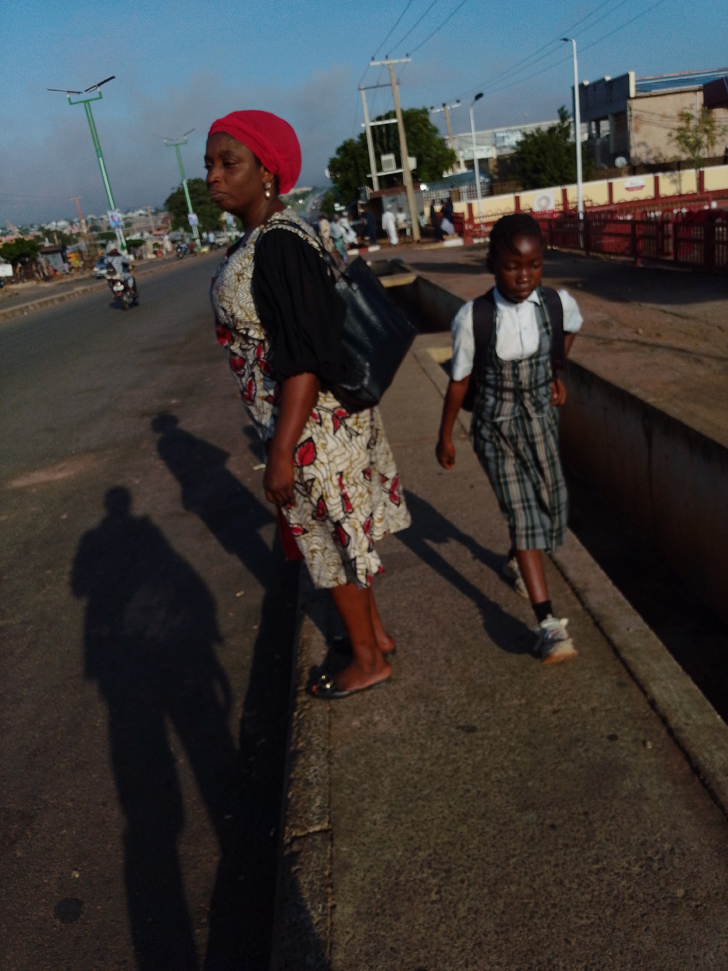 In Tumfure, Nigeria, a woman wearing a red headscarf and a patterned dress stands on a sidewalk beside a road. She is looking to her left, and her right hand is raised slightly. A young girl in a school uniform, consisting of a white shirt and a plaid jumper, walks in front of the woman, looking down. The girl carries a backpack. They are on a paved area with a curb. In the background, there is a road with traffic, including motorcycles, and buildings. The sky is clear and blue. The scene appears to be during daylight.