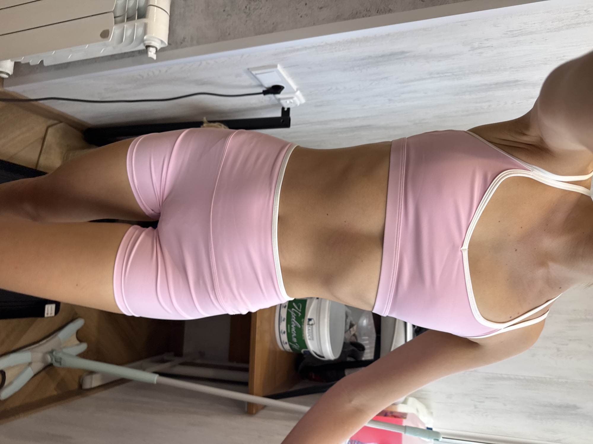 A person is standing and facing the camera, wearing a pink sports bra and pink shorts. The background includes a white wooden wall, a radiator, an electrical outlet, a wooden shelf with a white bucket, and a mop. The lighting appears to be natural and coming from the right.
