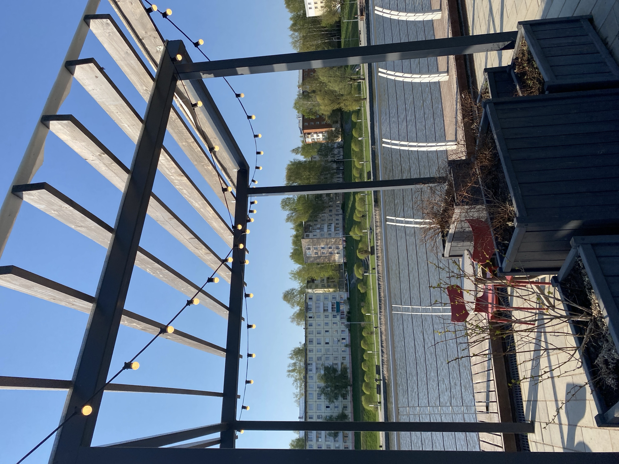 The image shows an outdoor seating area with a pergola structure overhead. Strings of lights are hung along the pergola. The seating area is adjacent to a body of water with a paved walkway running alongside it. Across the water, there is a row of apartment buildings visible, suggesting a residential area. The image is taken from the city of Votkinsk, Russia. The seating area contains two red chairs and is flanked by black planters. The ground is paved with grey stone. The scene is bathed in bright sunlight under a clear blue sky. There are no people visible in the image.
