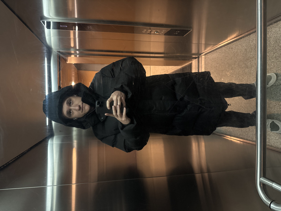 The image, oriented 90 degrees clockwise, captures an individual taking a full-body selfie within a modern elevator. The person is dressed in a dark, possibly black, hooded puffer coat with the hood up. They hold a smartphone in their right hand, positioned to take the photo, and have earphones in their ears. Their gaze is directed towards the camera with a neutral expression.
The elevator interior features highly reflective, polished metal walls that prominently mirror the individual and the surrounding environment. An elevator control panel with buttons and indicators, including a faintly visible 'F1' marking, is reflected on the upper section of one wall. Warm, artificial light sources illuminate the scene, casting reflections across the metallic surfaces and suggesting an indoor setting, possibly during the evening or night. The elevator's floor is a light tone, and beyond the open or transparent elevator doors, a similar light-colored floor or ground is visible. A metallic railing or door frame runs along the right side of the frame. This scene is situated in Warsaw, Poland.