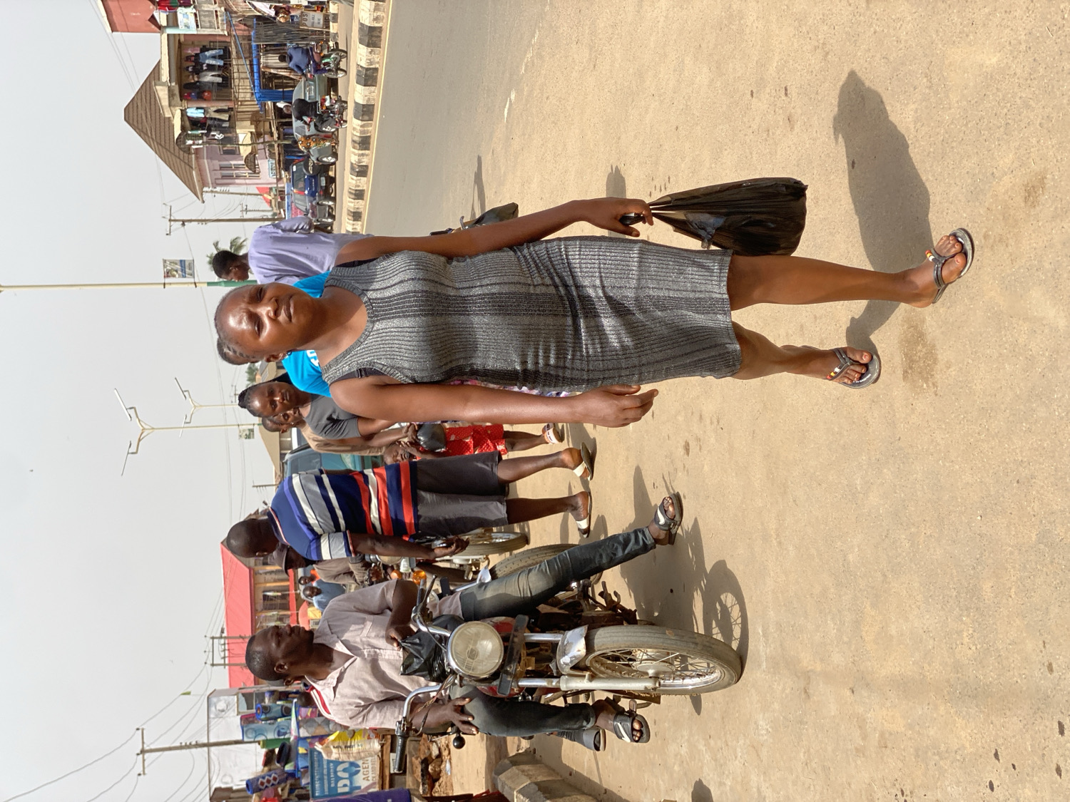 This media file captures a vibrant street scene, rotated 90 degrees clockwise, in what appears to be a commercial or market area of Kafanchan B, Nigeria, under bright daylight.
**Subjects:** The central figure is a woman in the foreground, wearing a fitted, shimmering grey sleeveless dress and silver-colored flip-flops. She holds a black plastic bag in her right hand and looks directly at the camera with a neutral expression. Behind her, several other individuals are visible, including a man in a light blue t-shirt and another in a blue, red, and white striped shirt. In the lower part of the frame, a man wearing a light-colored collared shirt and dark pants is seated on a motorcycle, looking to his left. Other pedestrians and figures are partially visible in the mid-ground and background, suggesting a busy environment.
**Objects:** Prominent objects include the woman's black plastic bag and the motorcycle in the foreground. In the background, there are various buildings that appear to house shops or stalls, some with awnings. Street poles, possibly for lighting or utilities, are also visible. The ground is a light, dusty brown.
**Activity:** The scene depicts daily life and movement. People are walking, standing, and riding motorcycles, indicative of routine transit and commerce. The main woman appears to be posing for the photograph.
**Notable details:** The time of day is clearly bright daytime, evidenced by the strong shadows cast by the subjects and the motorcycle. The sky, though largely overexposed, appears clear, suggesting sunny weather. The general atmosphere, architecture of the buildings, and presence of motorcycles as a mode of transport are characteristic of an urban or semi-urban setting in Nigeria.
**Visible text:** Small, unreadable text can be seen on signs and packaging near the shops in the background. On one blue sign, fragments like "AGEN" and "DA" are faintly discernible.