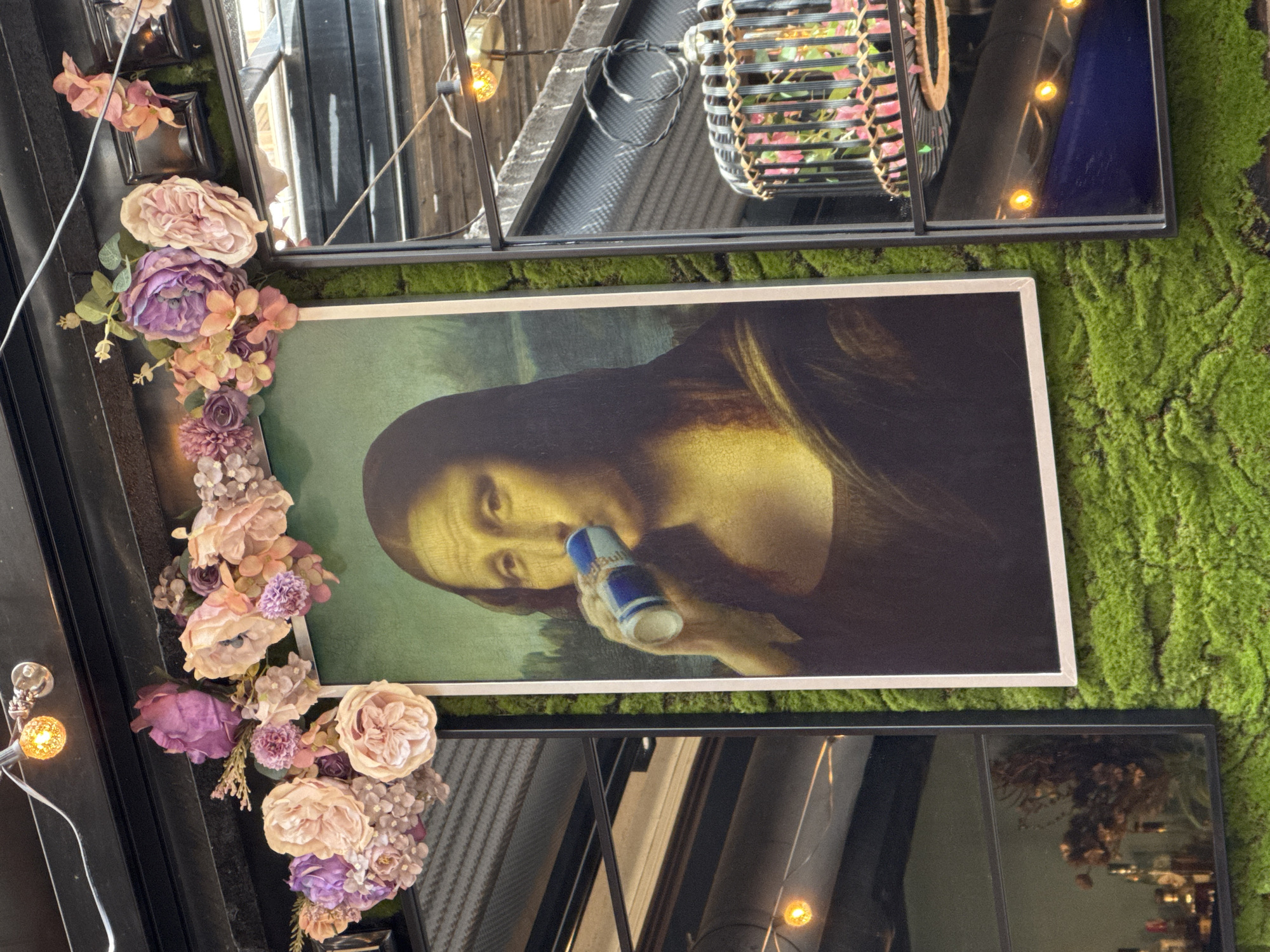 The image shows a framed print of the Mona Lisa drinking a can of Red Bull. The print is hung on a wall covered in green moss. The frame of the print is white. Surrounding the Mona Lisa print are a series of black-framed mirrors and floral decorations featuring pink and purple flowers. The scene appears to be indoors, perhaps in a cafe or restaurant in Pleven, Bulgaria. Small light bulbs can also be seen.