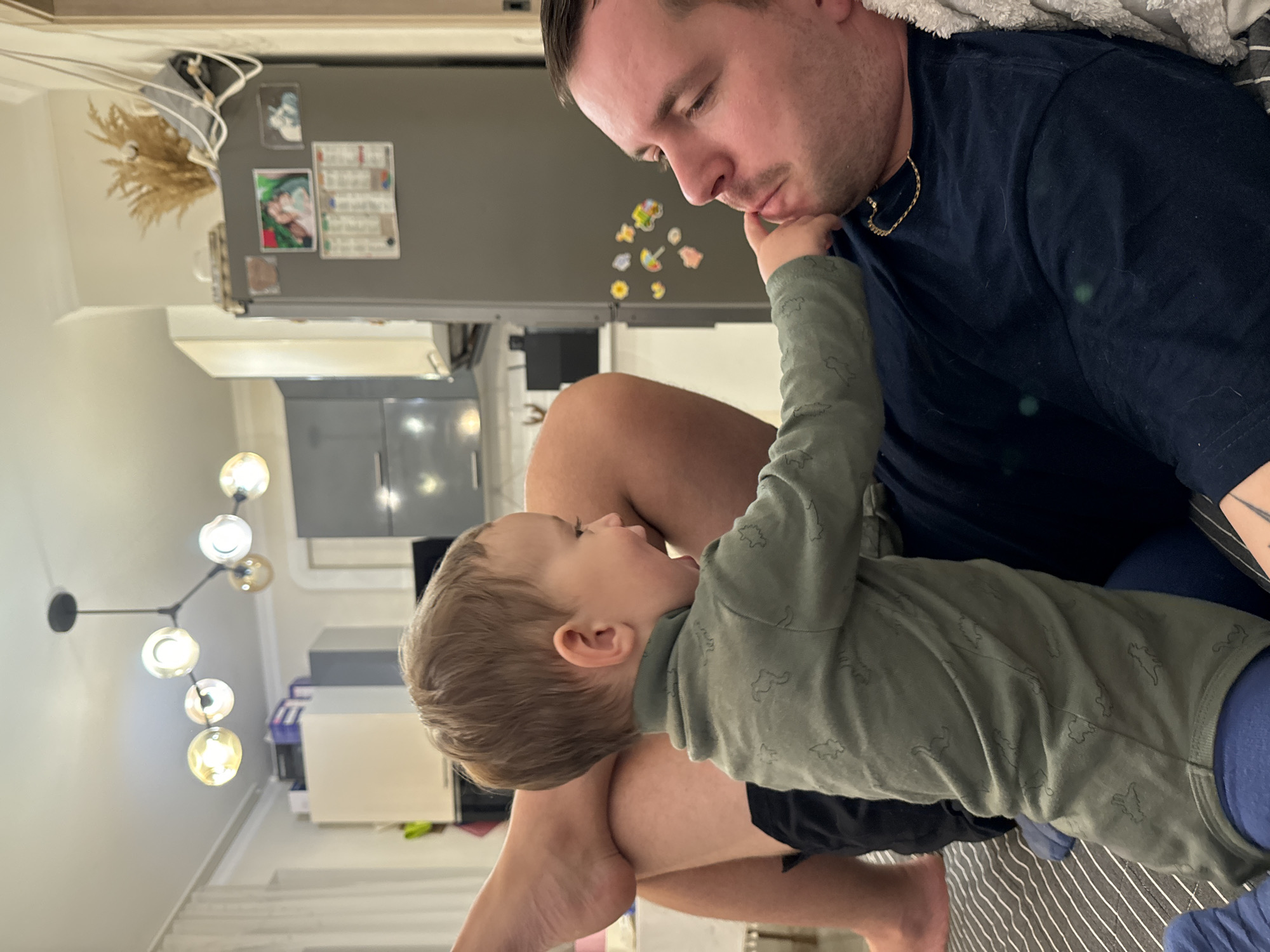 This indoor photograph captures a tender moment between an adult male and a young child in a domestic setting, likely a home in Odintsovo, Russia. The image is oriented vertically, but the room's elements, such as the refrigerator and light fixture, indicate the scene's true verticality.
The adult male, possibly a father figure, occupies the right side of the frame. He has short dark hair and light facial stubble, and is wearing a dark blue t-shirt with a gold chain. He is looking down gently at the child.
On the left, a young child, a toddler with light brown hair, is engaging with the man. The child wears a long-sleeved olive-green shirt patterned with faint outlines of what appear to be dinosaurs or small animals. The child's left hand is raised, softly touching the man's chin or jaw, creating a sense of connection and intimacy.
The background reveals elements of a home interior, possibly a kitchen or an open-plan living space. A large grey refrigerator, decorated with several magnets and small pictures, including one with unreadable text, is prominent behind the man. Above the refrigerator, a bundle of dried wheat or pampas grass is displayed. To the left, a modern chandelier with multiple glowing globe-shaped bulbs hangs, illuminating the scene. A striped blanket or sheet is visible beneath the subjects. The subdued indoor lighting suggests a comfortable, possibly evening, atmosphere, emphasizing the quiet, affectionate interaction between the man and child.