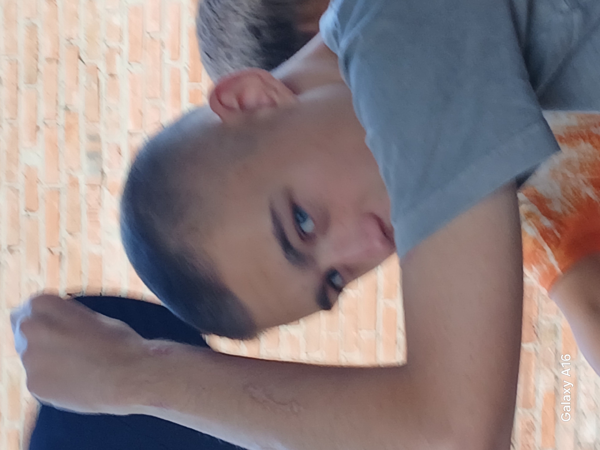 The image shows a close-up of a young person with light skin, short dark hair, and blue eyes. The person is wearing a gray t-shirt and has their left arm raised, with their fist clenched near their head. The background is a brick wall. There is also a hint of another person behind the main subject, with dark hair. The lighting suggests it is daytime, possibly outdoors. The bottom left corner of the image shows the text "Galaxy A16." The location is identified as Ustyvytsia, Ukraine, implying the scene is within or near this Ukrainian settlement.