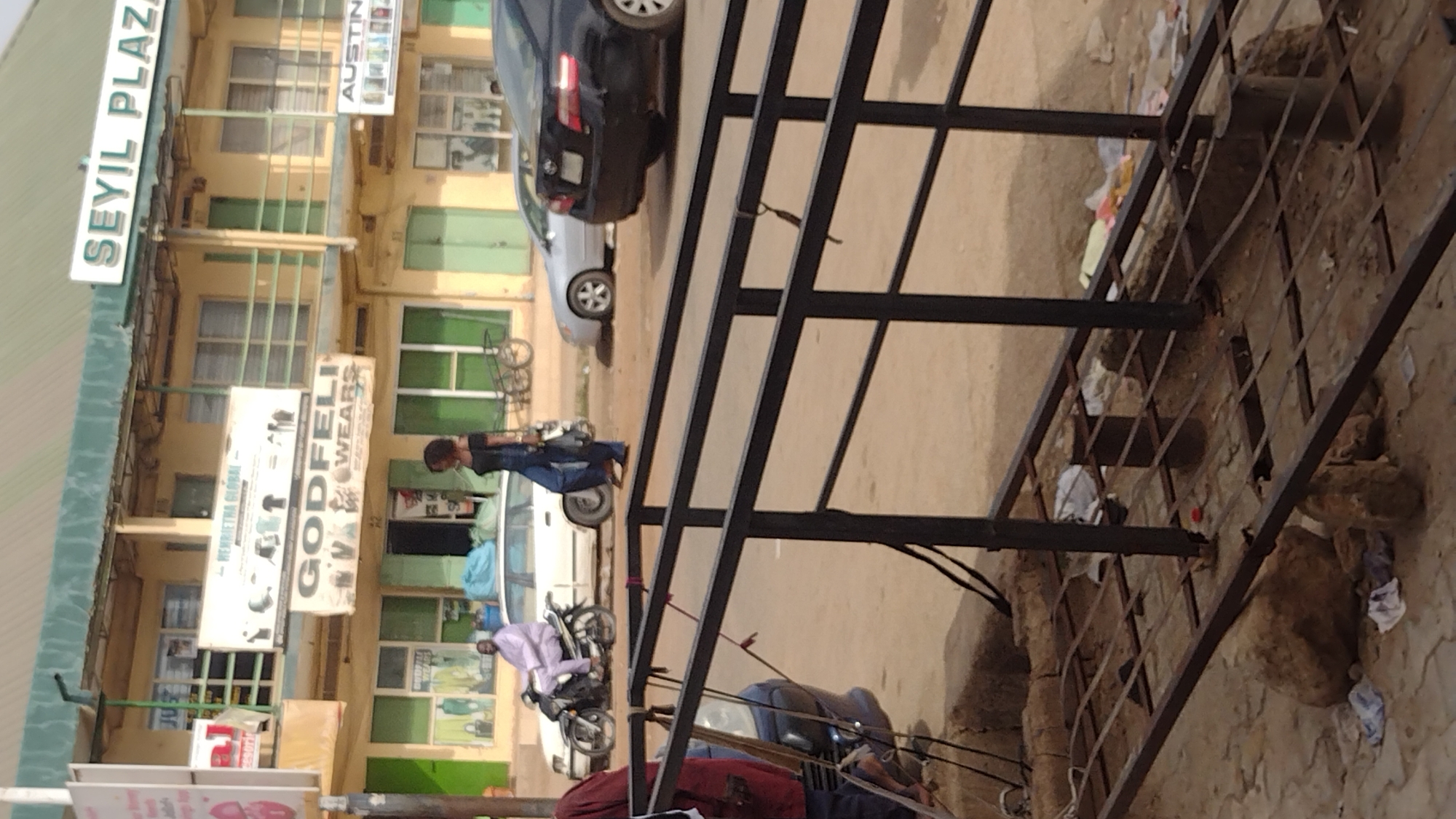 The image depicts a street scene in Jos, Nigeria, featuring a building labeled "Seyil Plaza." The building is multi-storied with multiple businesses advertised on the exterior, including "Godfeli Wears" and "Austin." Several cars and motorcycles are parked along the street. People are walking and standing nearby. A black metal structure dominates the foreground. The overall environment appears to be urban, with visible litter and debris scattered on the ground.