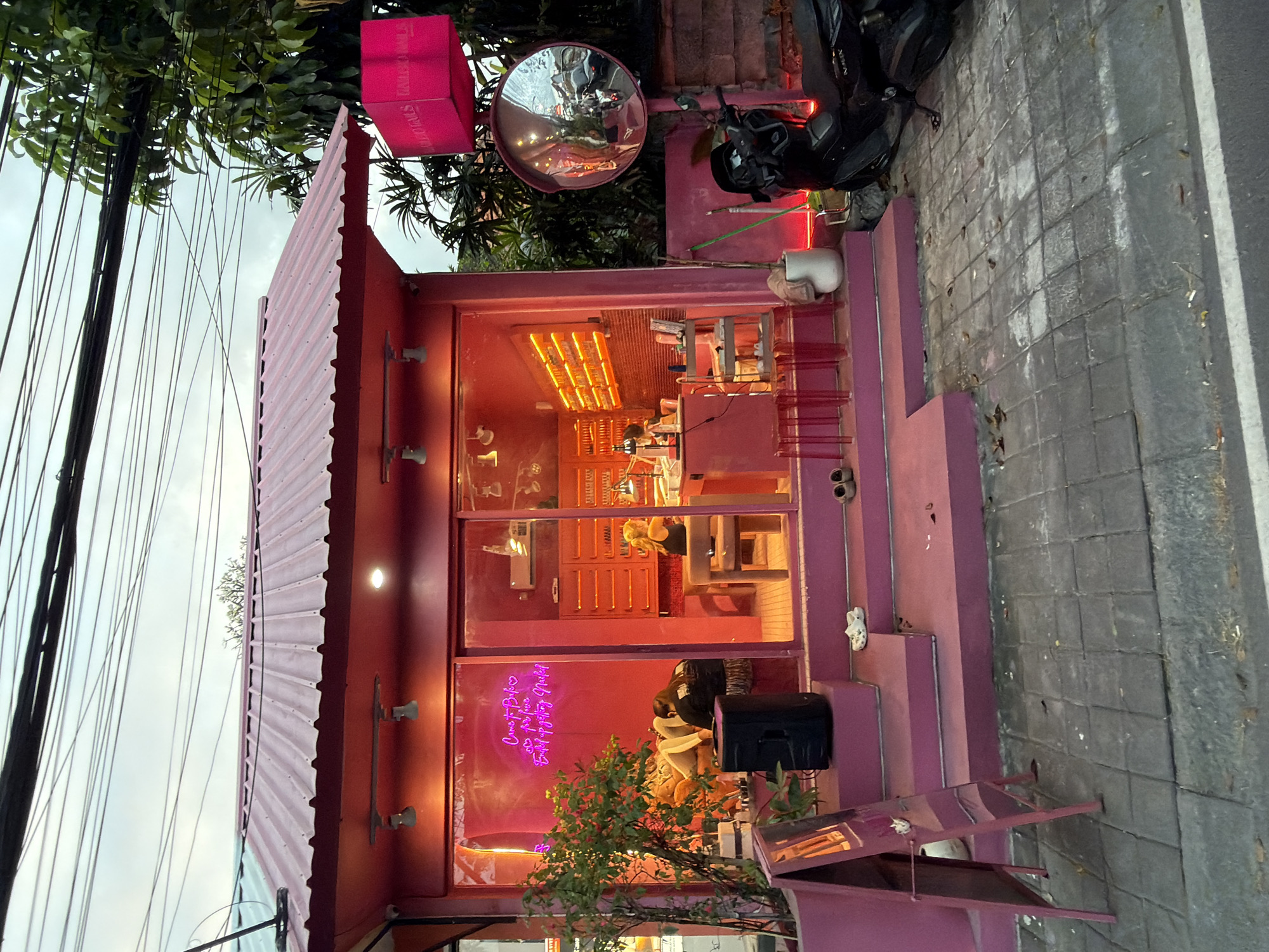 The image captures a vibrant pink building in North Kuta, Indonesia, likely a nail salon, as indicated by the neon sign inside reading "Come to Bubu's & Get your Nice, Bright & Shiny Nails." The storefront features large glass windows revealing the interior, where two women are visible. One woman is reclined on a lounge chair, seemingly receiving a treatment, while another sits at a table, engaged in nail work. The interior is illuminated by bright pink and orange lighting, with shelves stocked with nail polish bottles.
Outside, the building has a tiered pink facade with steps leading up to the entrance. A black speaker is placed on the steps, and several pairs of sandals are scattered around. A pink A-frame sign stands to the left. To the right of the building, a large, convex mirror is mounted on a pole, reflecting the street scene with passing vehicles. A black scooter is parked next to the mirror. The building is situated on a paved walkway, with overgrown trees and vegetation visible behind it. Overhead, a tangle of electrical wires crisscrosses the sky, typical of urban environments in Indonesia. The overall scene suggests a lively and colorful commercial establishment.