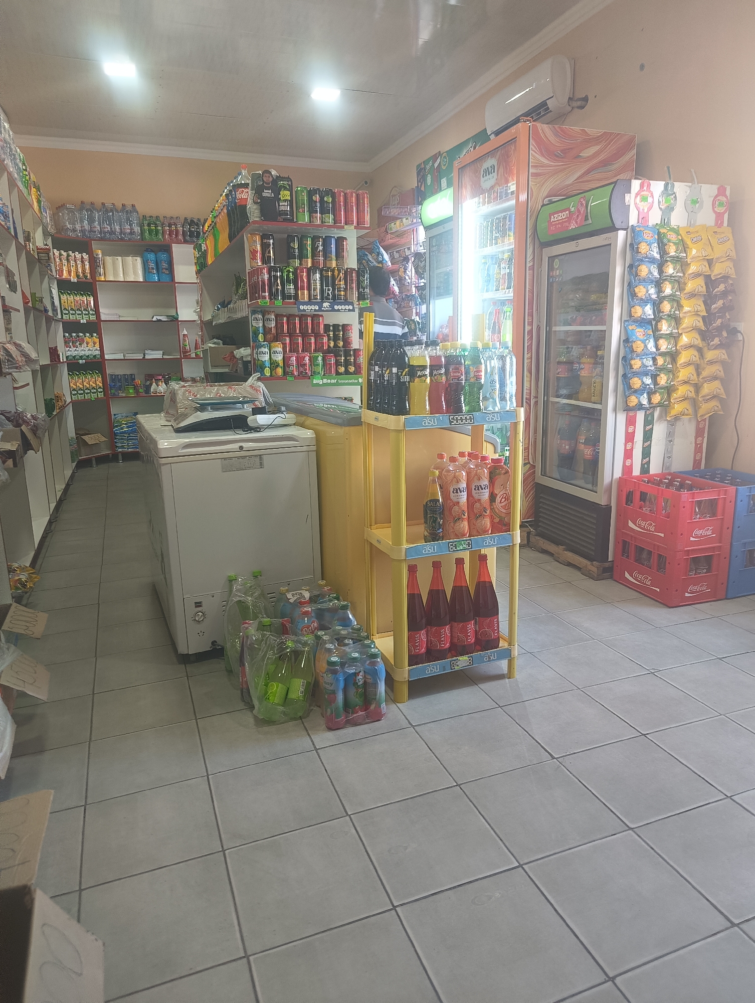 The image depicts the interior of a small convenience store, possibly in Bulungur, Uzbekistan. The store features several shelves stocked with various beverages and snacks. On the left, shelves display rows of bottled water, sodas, and juices. In the center, a white chest freezer is situated, with a scale on top. Next to the freezer, a yellow shelving unit holds more bottled drinks, including some with orange and red labels. The right side of the store has refrigerated display cases filled with beverages and a rack of packaged snacks. Several red and blue Coca-Cola crates are stacked on the floor in the background. The floor is tiled, and fluorescent lights illuminate the space. No people are clearly visible in the image, but the overall impression is one of a well-stocked retail environment.