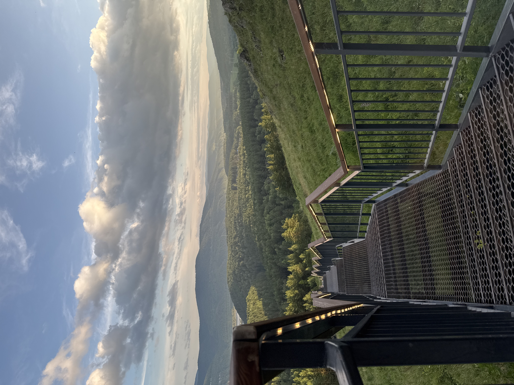 This is a vertical, eye-level, medium shot of a scenic overlook with a metal staircase descending into a lush, forested valley under a dramatic sky. The primary subjects are the staircase, the valley landscape, and the sky.
The metal staircase, made of dark gray metal with wooden handrails, dominates the right side of the frame. Its steps are made of perforated metal, and the railing has a simple, modern design. The staircase appears to be part of a viewing platform, with railings and a wooden top edge visible.
Beyond the staircase, the scene unfolds into a vast, rolling valley covered in dense, green forest. The trees are a deep, vibrant green, suggesting a warm season. In the distance, several layers of mountains fade into a soft, hazy blue, indicating depth and distance. The sky is a mix of clear blue and large, puffy white and gray clouds, illuminated by what appears to be the warm light of either sunrise or sunset. The clouds are dramatic and textured, adding a sense of grandeur to the scene.
The overall mood is one of awe and tranquility, inviting the viewer to imagine descending the stairs and exploring the natural beauty below. The time of day is likely early morning or late afternoon, given the soft, golden light on the clouds and the long shadows that would be cast. The location is suggested to be Krasnoyarsk, Russia, a region known for its stunning natural landscapes. There is no visible text in the image.