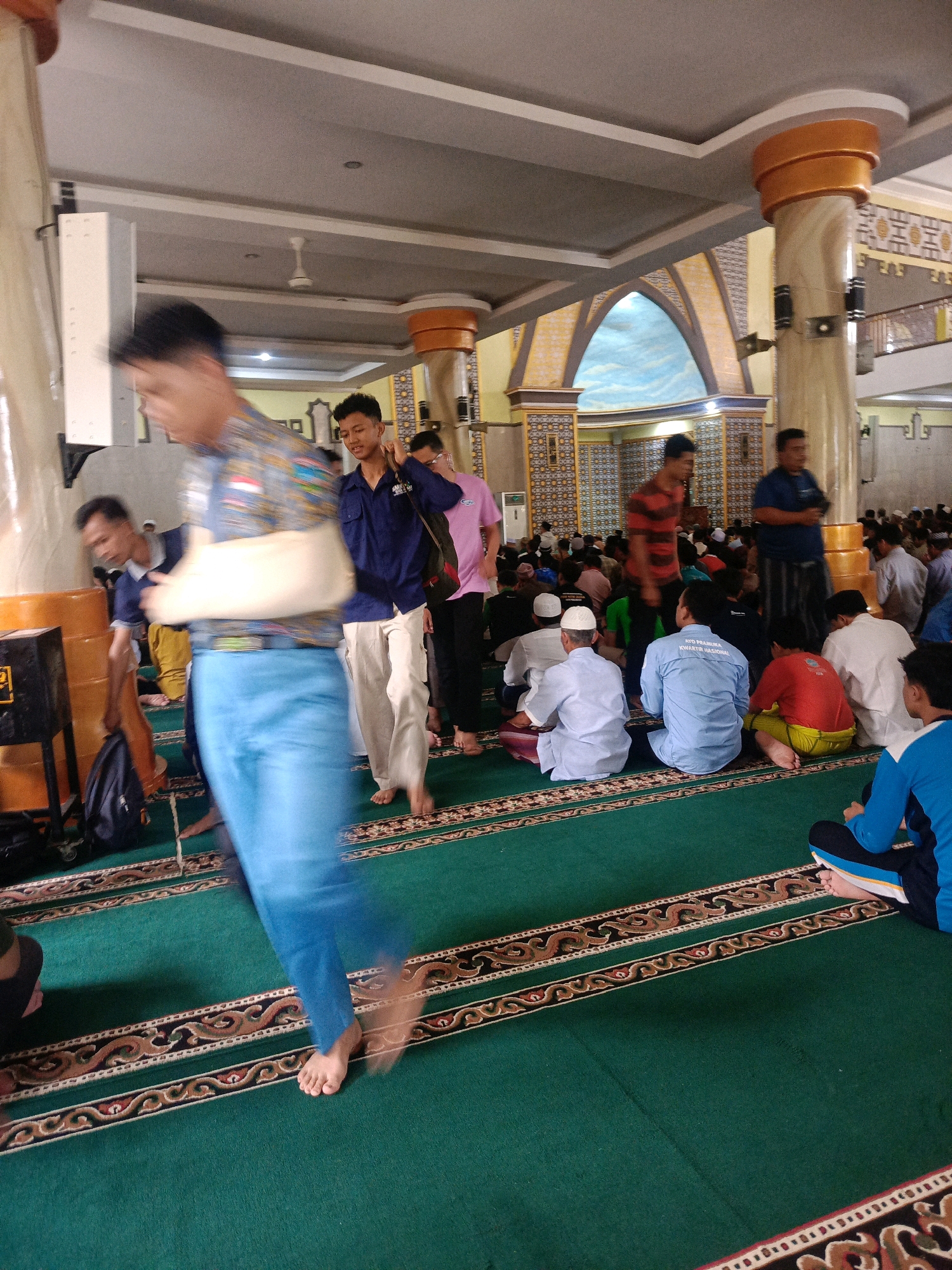 The image depicts the interior of a mosque, likely located in Magetan, Indonesia, given the context. The flooring is covered with a green carpet featuring decorative borders. Numerous individuals, predominantly men, are present, some seated on the carpet, while others are standing or walking.
In the foreground, a person in motion is captured with a blurred effect, wearing blue trousers and a patterned shirt with a cast on their left arm. To their right and slightly behind, another individual in a dark blue shirt is visible. Further into the scene, many people are seated, facing in various directions, some wearing white caps or peci. Several individuals are standing, including one in a red and black striped shirt and another in a blue shirt.
The mosque's architecture is visible in the background, with large pillars adorned with ornate designs and arched structures with painted murals depicting a sky scene. The lighting suggests an indoor environment. The overall impression is of a gathering within a mosque, possibly for prayer or a religious event.