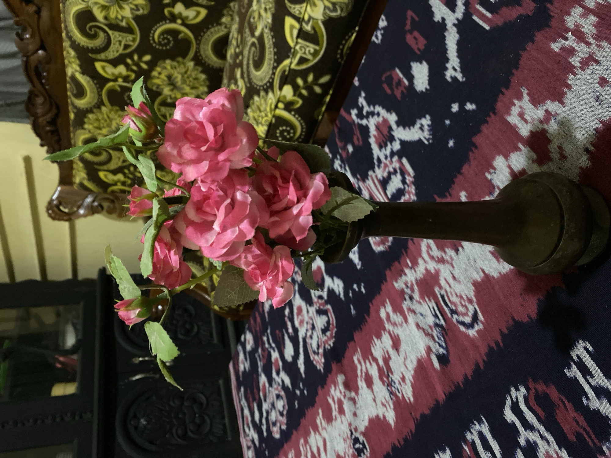 The image depicts an indoor still life arrangement. In the foreground, a dark brown vase holds a bouquet of artificial pink roses with green leaves. The vase is placed on a table covered with a patterned textile that features dark blue, pink, and white designs. In the background, a dark wooden cabinet with decorative carvings is visible to the left. To the right, a portion of a patterned sofa with dark brown and green floral motifs is shown. The overall lighting suggests an indoor setting, possibly a living room. The specific location within Gumelar Kidul, Indonesia cannot be determined from the image alone.