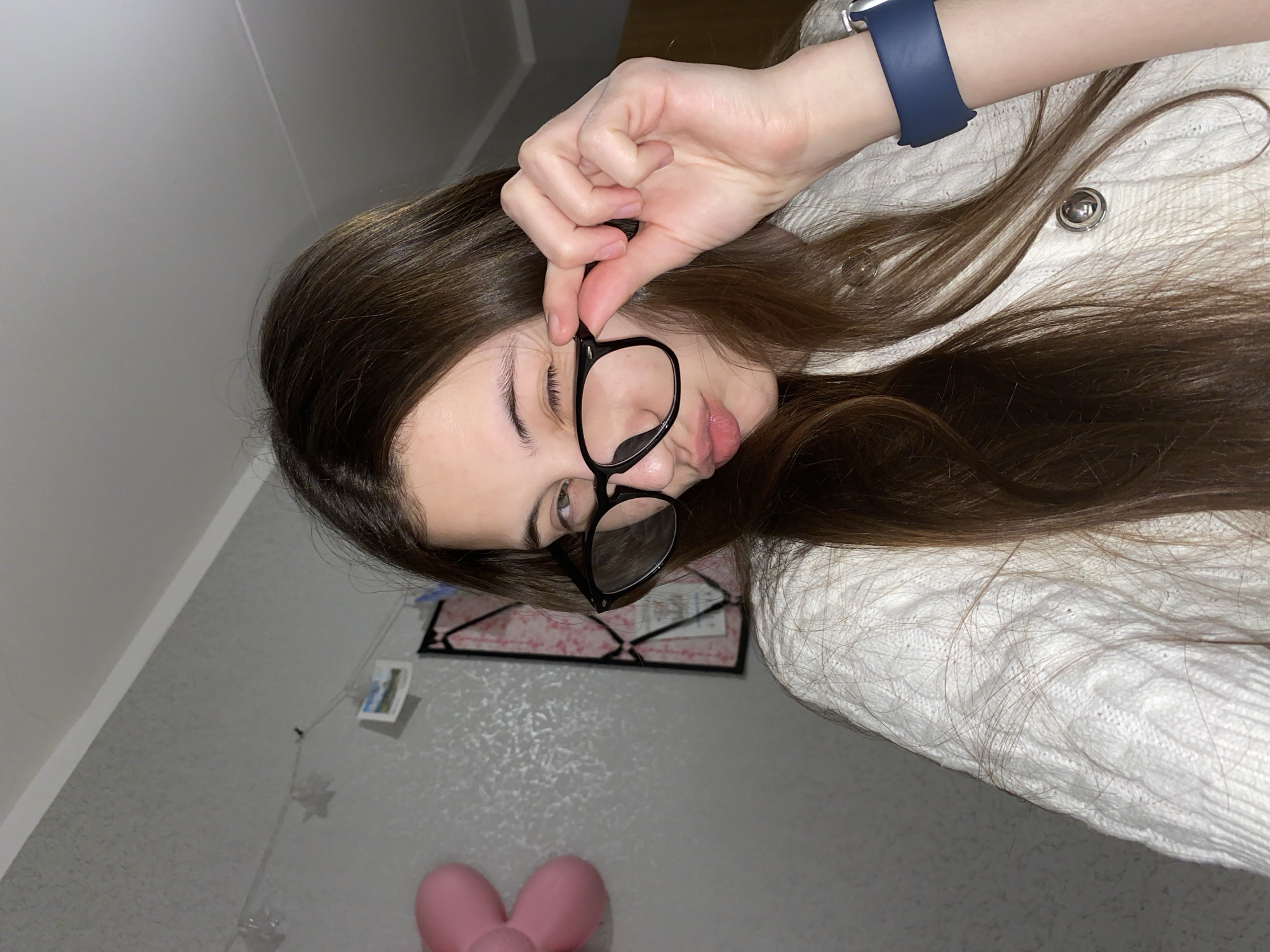 A young woman with long, dark brown hair is depicted in a close-up, overhead shot, appearing to be lying down and looking directly at the camera. She wears black-framed glasses, which she holds slightly down her nose with her right hand, peering over them with a curious or playful expression. On her left wrist, she wears a navy blue Apple Watch. She is dressed in a white, textured cable-knit sweater, with some strands of her hair resting upon it.
The background consists of light grey walls, which show a subtle texture and a faint line near the top left corner. A string adorned with small, indistinct items, possibly photos or decorative elements, hangs vertically on the wall behind her. Partially visible below her head is a framed object, possibly artwork or a document, featuring a pink patterned surface. In the bottom center of the frame, a bright pink, bow-shaped object rests on the floor or surface. No discernible text is visible in the image. The location context for this image is Votkinsk, Russia.