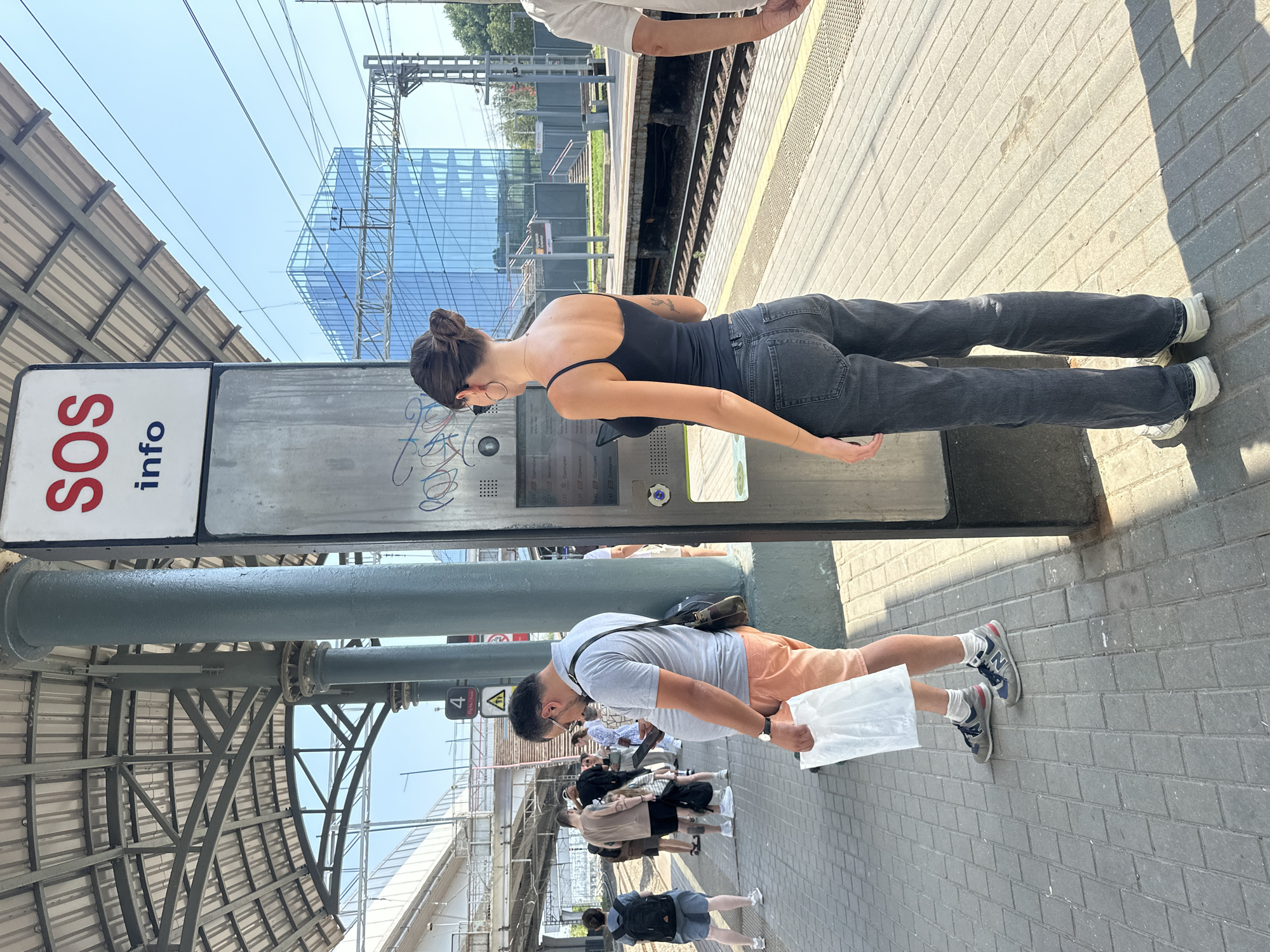 The image shows a sunny day at a train station platform in Khimki, Russia. A woman with dark hair in a bun, wearing a black tank top and dark jeans, stands facing an SOS information kiosk and appears to be looking at a phone. A man in a grey polo shirt and orange shorts, holding a white plastic bag, stands to her left, also looking at his phone. Several other people are visible on the platform in the background, as well as train tracks and a modern glass building in the distance. The platform is paved with grey bricks.