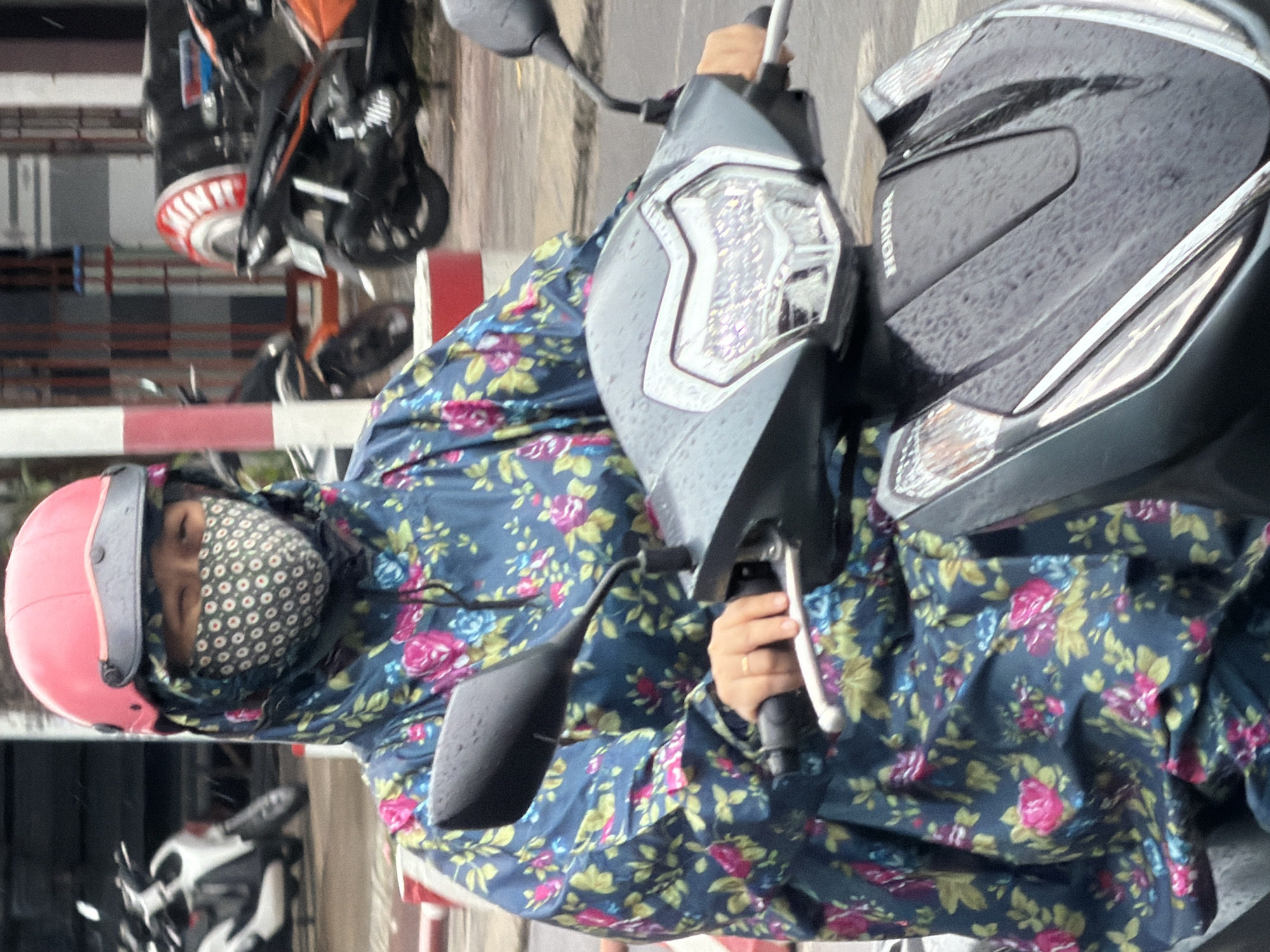 A person wearing a pink helmet and a floral print raincoat is riding a dark grey scooter on a wet street. The person's face is covered by a patterned fabric mask and the hood of the raincoat. The scooter has a Honda logo on the front. Other scooters and buildings are visible in the background, suggesting an urban environment. The scene appears to be in Nha Trang, Vietnam, given the context of the provided image.