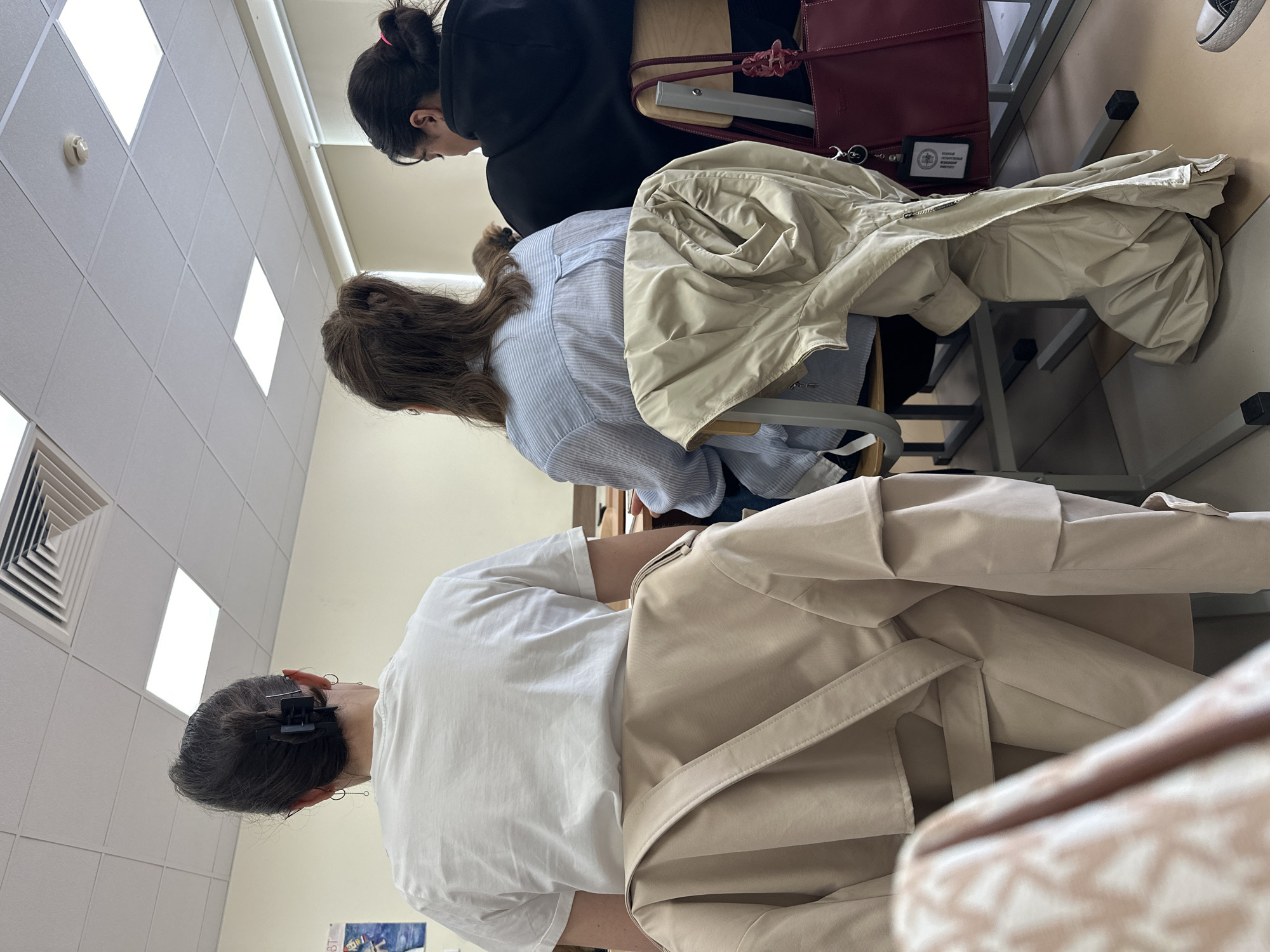 The image depicts an indoor scene, likely a classroom or lecture hall in Kazan, Russia, showing three individuals, presumably students, seated at desks. They are viewed from behind and the side.
In the foreground, a person with dark hair tied back with a black claw clip wears a white t-shirt. A light beige/khaki trench coat or jacket is draped over their chair. The middle individual has long, wavy brown hair and wears a light blue striped button-down shirt. Another beige jacket is visible on the chair or desk in front of them. The person furthest back has dark hair tied up with a pink scrunchie and is wearing a black hoodie. A dark red handbag rests on their desk, alongside what appears to be an open notebook or folder.
A notable detail is a small black card or badge on the desk near the red handbag, partially displaying the Cyrillic text "РУССКИЙ ГОСУДАРСТВЕННЫЙ" (Russian State), confirming the setting as a Russian educational institution.
The ceiling features white square acoustic tiles with bright rectangular light panels and a white square ventilation grille. A white fire alarm or smoke detector is also mounted on the ceiling. The floor appears to be tiled in light, neutral colors. The individuals are engaged in an activity typical of a class or study session, sitting at their respective stations. No specific interactions or emotions are discernible as only their backs and sides are visible. The lighting is artificial, suggesting an indoor environment.