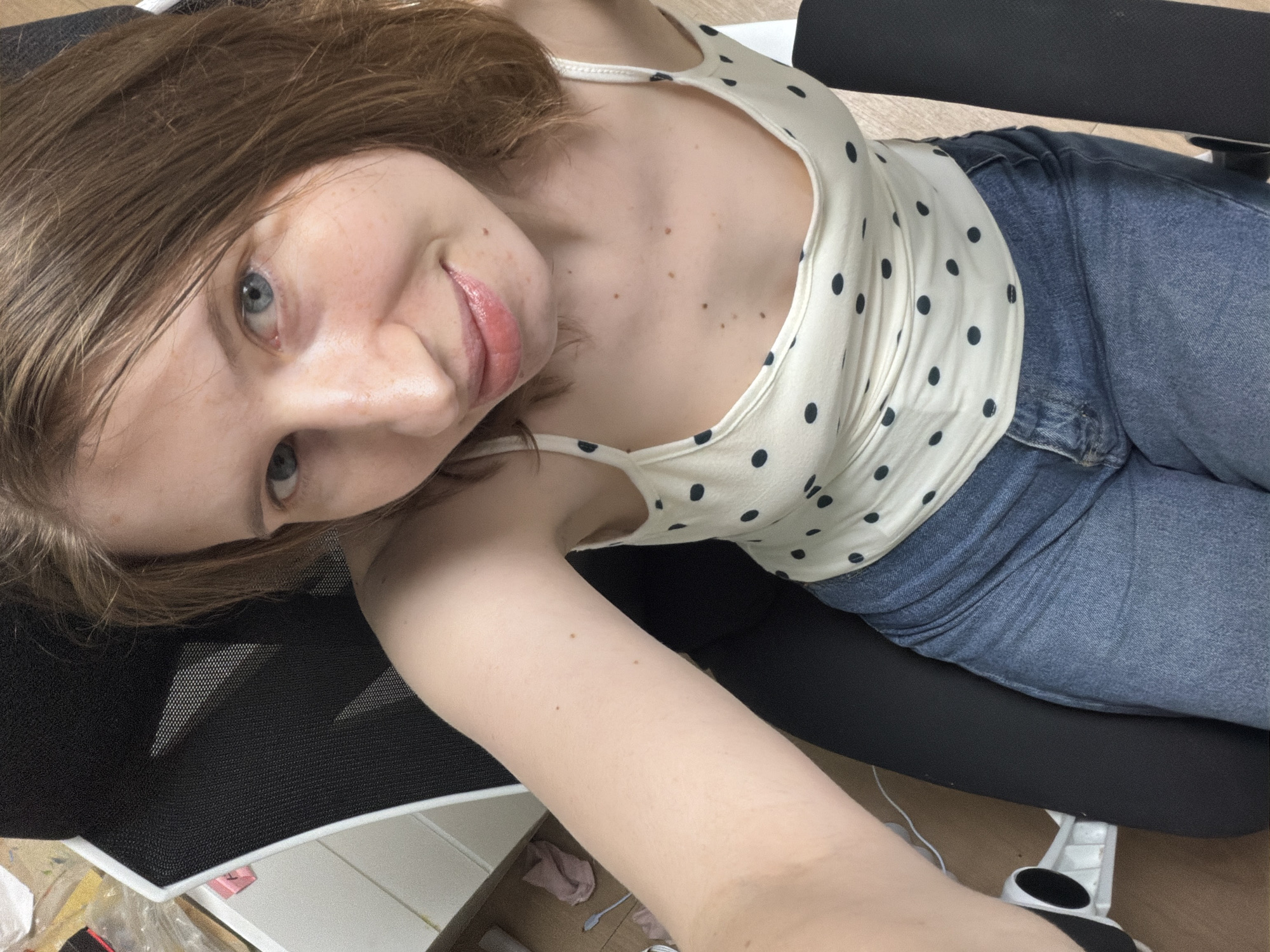 A fair-skinned woman with medium-brown hair and blue eyes is photographed from a slightly elevated, selfie-style angle. She is seated in a black office chair with a mesh back and white trim. She is wearing a white camisole with black polka dots and blue denim jeans. Her left arm is extended upwards and to the left, capturing the image. The background shows a desk or shelf with various items, including a pink cloth, and a white piece of office equipment with black accents, likely part of the chair. The overall lighting is bright, suggesting an indoor setting. No specific location in Dongducheon-si, South Korea is identifiable from the visual cues.