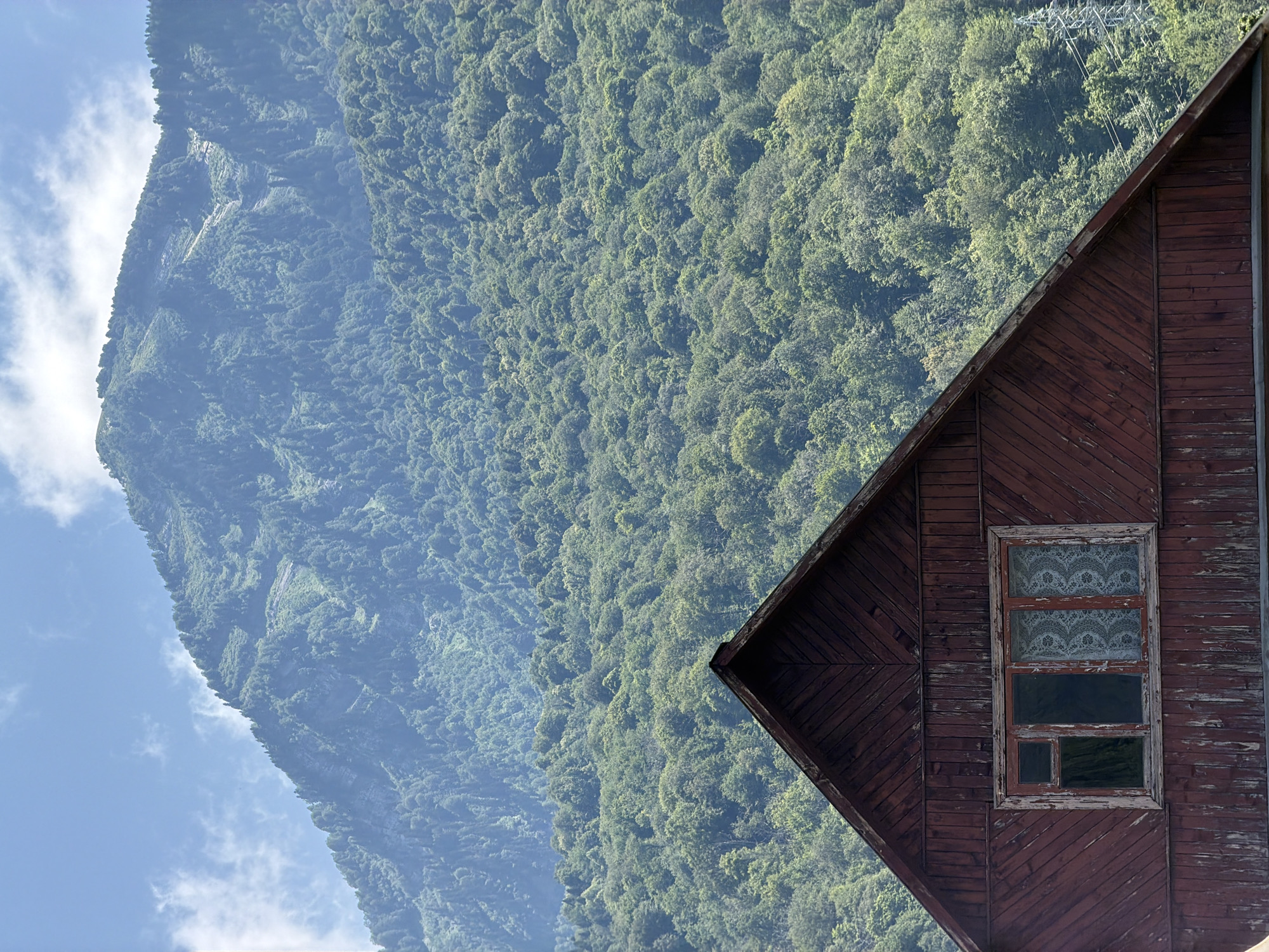 An aerial perspective of a wooden house gable in Krasnaya Polyana, Russia, reveals a lush, green mountainside dotted with trees under a hazy, blue sky with scattered white clouds. The house's roof is dark red and weathered, with visible wood grain and signs of age. A multi-paned window is set into the gable, with one pane showing a reflection of dark trees, another appearing dark and empty, and the remaining panes covered by white lace curtains. The mountainside, a dense forest, stretches upwards into the distance, its contours softened by a light atmospheric haze. A distant electrical transmission tower can be seen to the right, partially obscured by the trees.