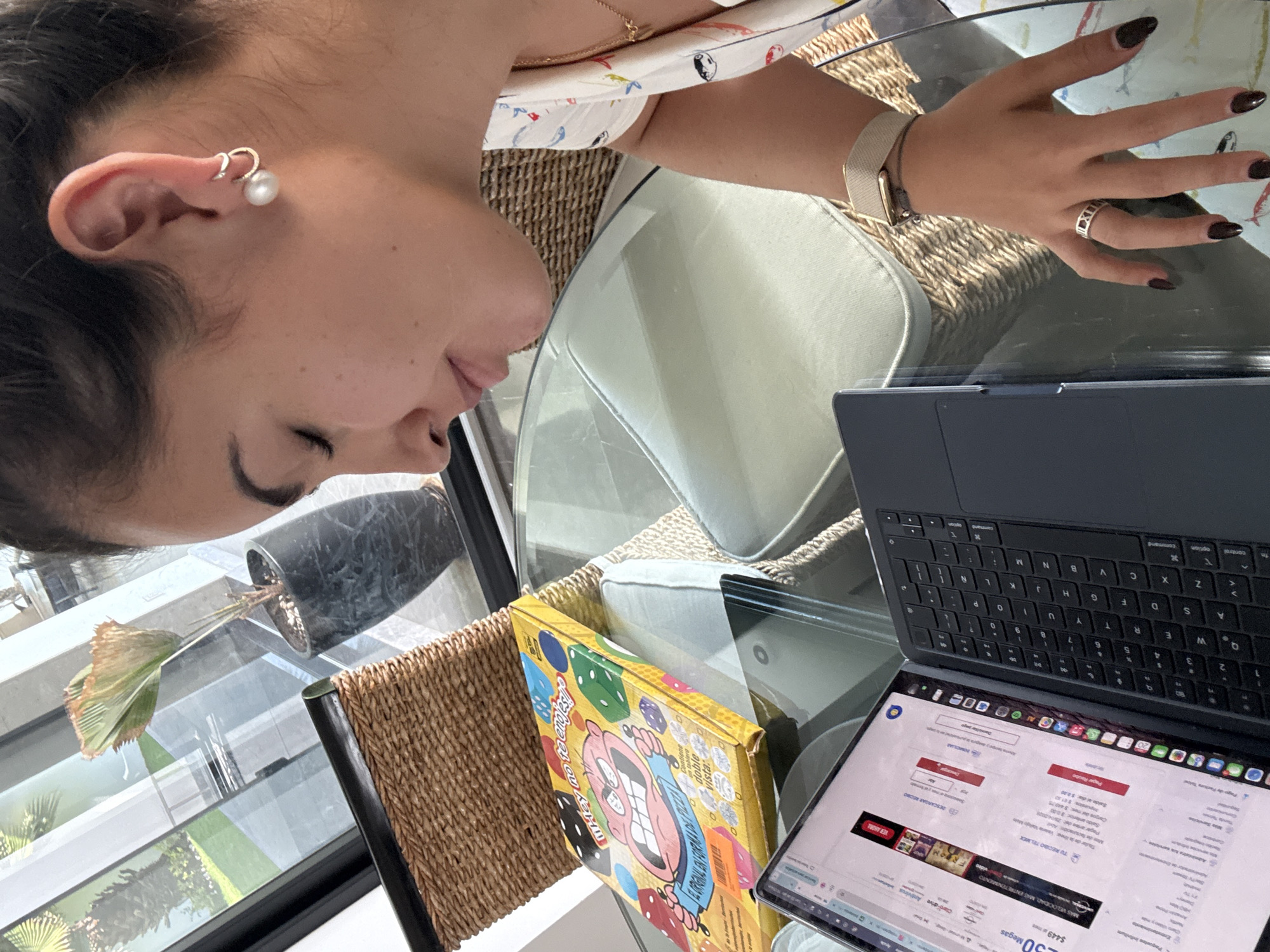 The image captures a close-up, overhead view of a person seated at a glass table, engaged with a laptop. The setting appears to be indoors, next to a large window or balcony, indicated by the reflection of green foliage, outdoor structures, and bright natural light suggesting daytime.
The primary subject is a person, likely female, whose head and right arm are visible from a profile angle. She has dark hair tied back and wears a white top with colorful, abstract patterns. Her right ear features a gold-toned cuff earring with a prominent white pearl. On her right wrist, she wears a light-colored Apple Watch. Her hand rests near the laptop, showcasing dark nail polish and a silver-toned ring with Roman numerals (specifically "XII" is visible). Her expression is neutral, seemingly focused.
On the glass table, a dark gray laptop with a matching detachable keyboard is open. The laptop screen displays a webpage, predominantly in Spanish, which appears to be related to a telecommunications bill or service. Visible text includes "TU RECIBO TELMEX" (Your TELMEX bill), "MAS VELOCIDAD, MAS ENTRETENIMIENTO" (More speed, more entertainment), "Claro-drive", "$449", and "30 Megas".
Next to the laptop, a bright yellow board game box is partially visible. The box features a cartoonish face with dice illustrations. Visible text on the box includes "Whey note Choles" (as per OCR, likely a stylized game title) and "Contenido de doble vista" (Double-sided content).
In the background, behind the glass table and through the window, a potted plant with large, somewhat withered leaves is visible. The reflection on the glass shows more distant buildings and lush greenery, providing context to the location, San Bernardino Tlaxcalancingo, Mexico, as supported by the Spanish text on the laptop screen and the local company names. The person is seated in a chair with a wicker back.