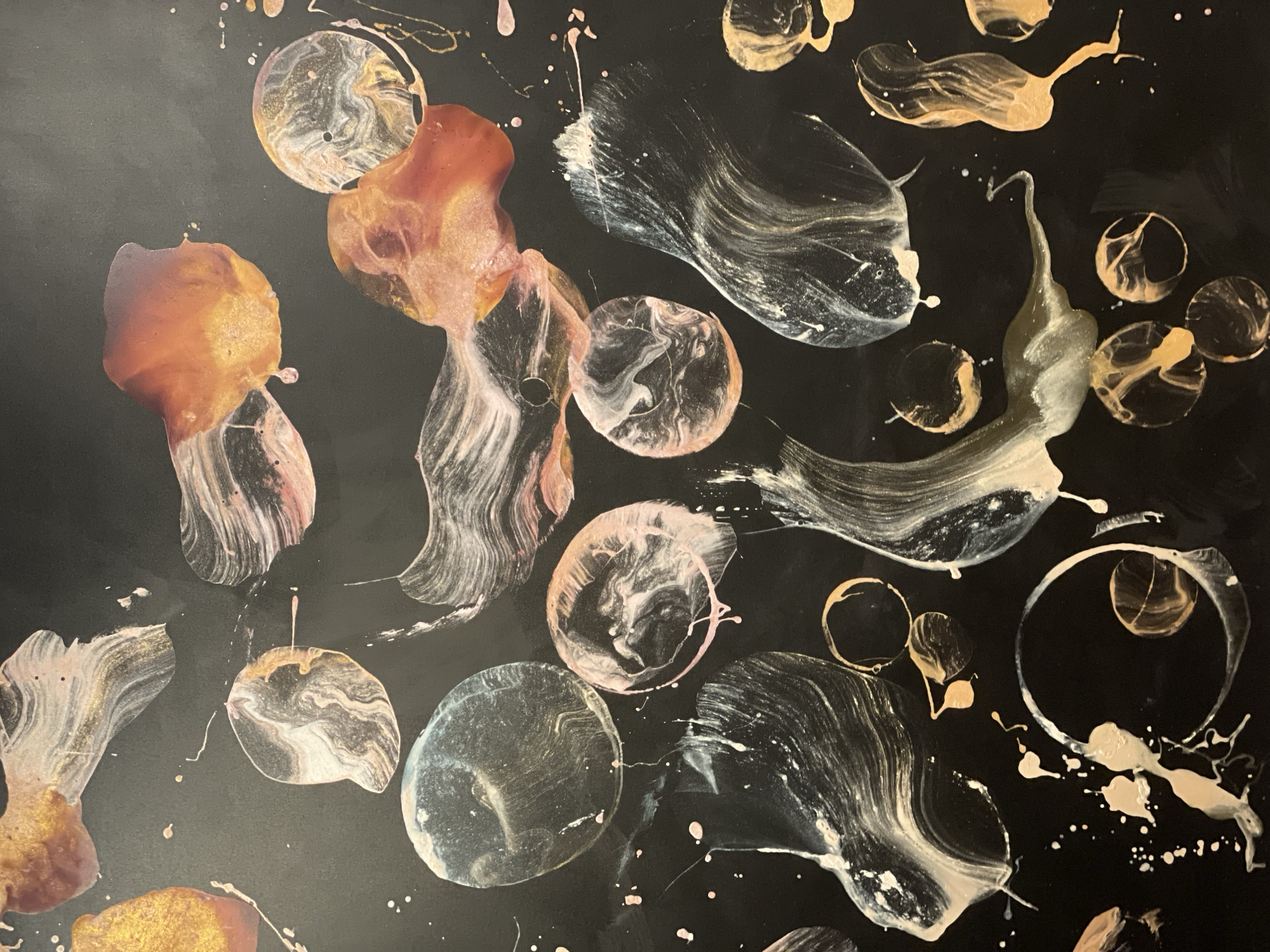 This is a vibrant, abstract close-up image of a fluid art painting. The dominant background is a glossy, deep black, providing a strong contrast to the colorful, organic shapes and patterns. The artwork features an array of swirling, dripped, and splashed forms in metallic gold, copper, white, and pale pink, with some hints of silver or light blue.
The shapes vary from distinct circles and rings, some appearing like bubbles or cells, to more amorphous, flowing designs that suggest movement and fluidity. Within these shapes, the colors are often swirled together, creating intricate marble-like textures and iridescent effects, particularly with the metallic paints which catch the light. Numerous small splatters and drips of paint, primarily white and pink, are scattered across the black surface, adding to the dynamic composition.
The overall impression is one of shimmering liquids in motion. The glossy finish suggests a resin coating or a similar technique that enhances the depth and sheen of the colors. There are no people, recognizable objects beyond the paint itself, or visible text in the image. No specific time of day, weather, or discernible location cues are present in the artwork itself.