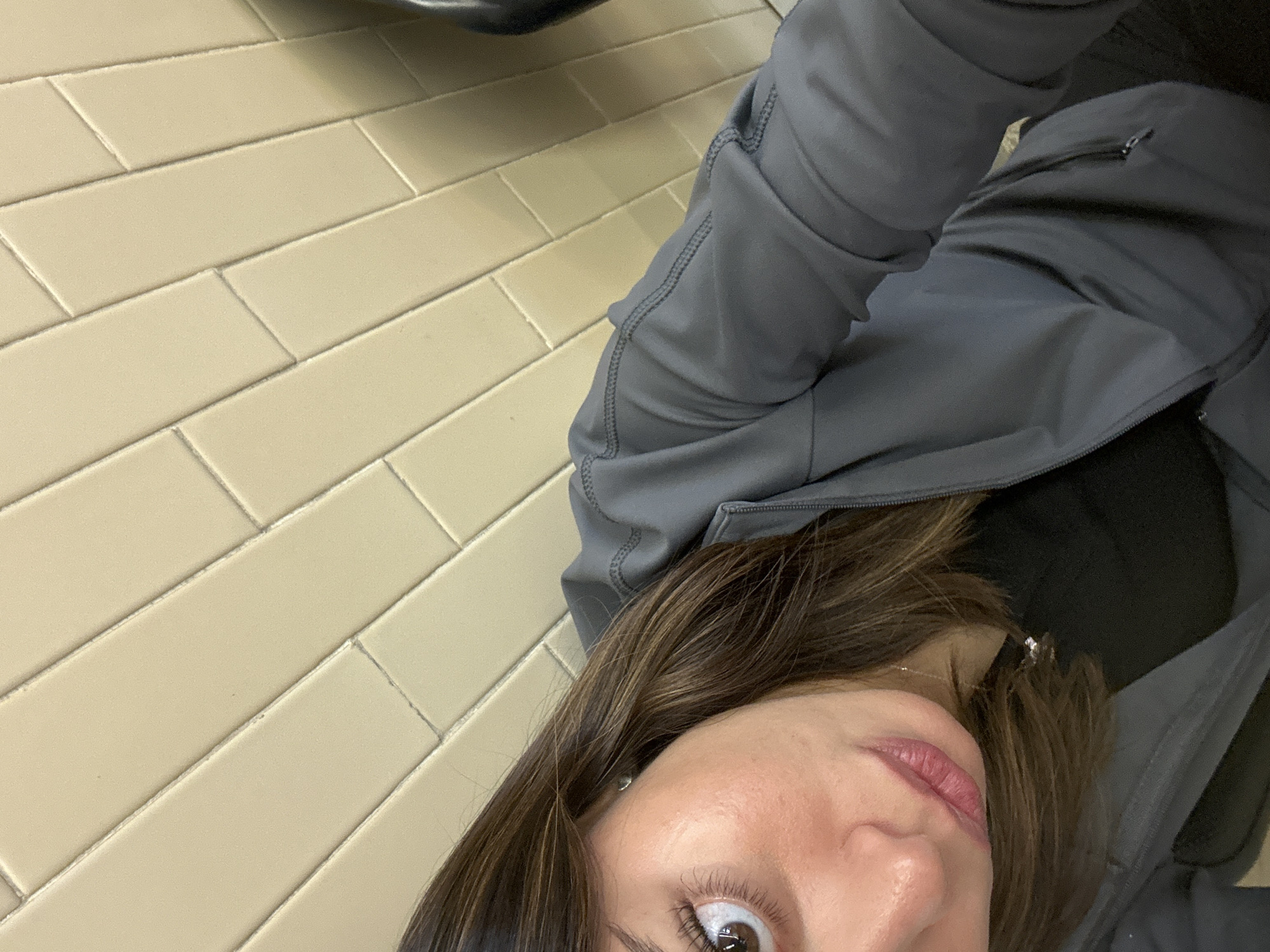 The image presents a unique, tilted, and angled close-up shot, appearing to be a selfie taken from a first-person perspective looking downwards. The primary subject is a person, whose upper body and a significant portion of their face are visible.
The person has long, medium-brown hair that frames the right side of their face. They are wearing a dark grey or charcoal-colored jacket, possibly made of a soft-shell or athletic material, with visible seams and stitching, particularly on the sleeves. A zippered pocket is visible on the upper arm of the jacket. Beneath the jacket, a hint of a black top can be seen.
Facial features include a wide-open dark brown eye with visible eyelashes, and slightly parted pink lips with a subtle sheen. The expression is neutral, with the eye looking directly towards the camera. A small, light-colored stud earring is visible in the ear, partially obscured by hair, and a thin silver-colored necklace is also visible near the collarbone.
The background is dominated by a light beige or cream-colored tiled floor. The rectangular tiles are laid in a staggered or brick-like pattern, with light-colored grout lines clearly visible between them. The strong diagonal lines of the tiles are a result of the extreme camera angle. The setting appears to be indoors, indicated by the tiled floor. There is no visible text or specific identifying location cues. The lighting seems even, possibly from overhead.