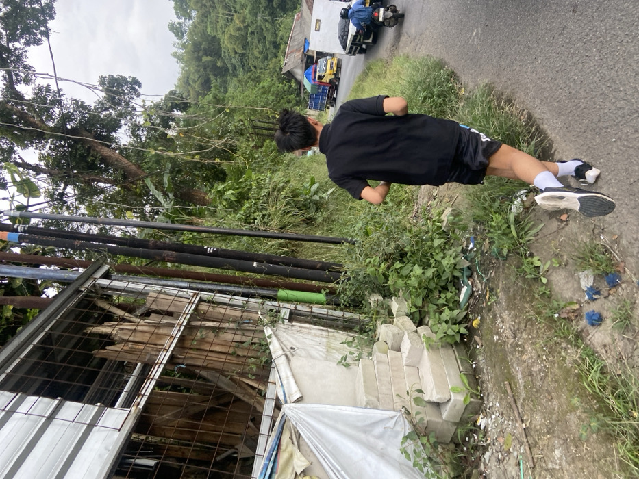A young man is running down a paved road in Cipatat, Indonesia, located at latitude -6.792 and longitude 107.434. The road is lined by lush green foliage and a wooden and metal structure on the left side. The structure is partially obscured by a white tarp. In front of the structure is a concrete staircase with a series of steps. There are several blue pieces of debris lying on the ground in front of the steps. The man is wearing a black t-shirt and black shorts. He is wearing white socks and white and black running shoes. His dark hair is pulled back in a ponytail. The road appears to be empty, though in the background a white truck and a red pickup truck can be seen driving away. The man is running to the left of the frame, with his right foot extended in a stride.