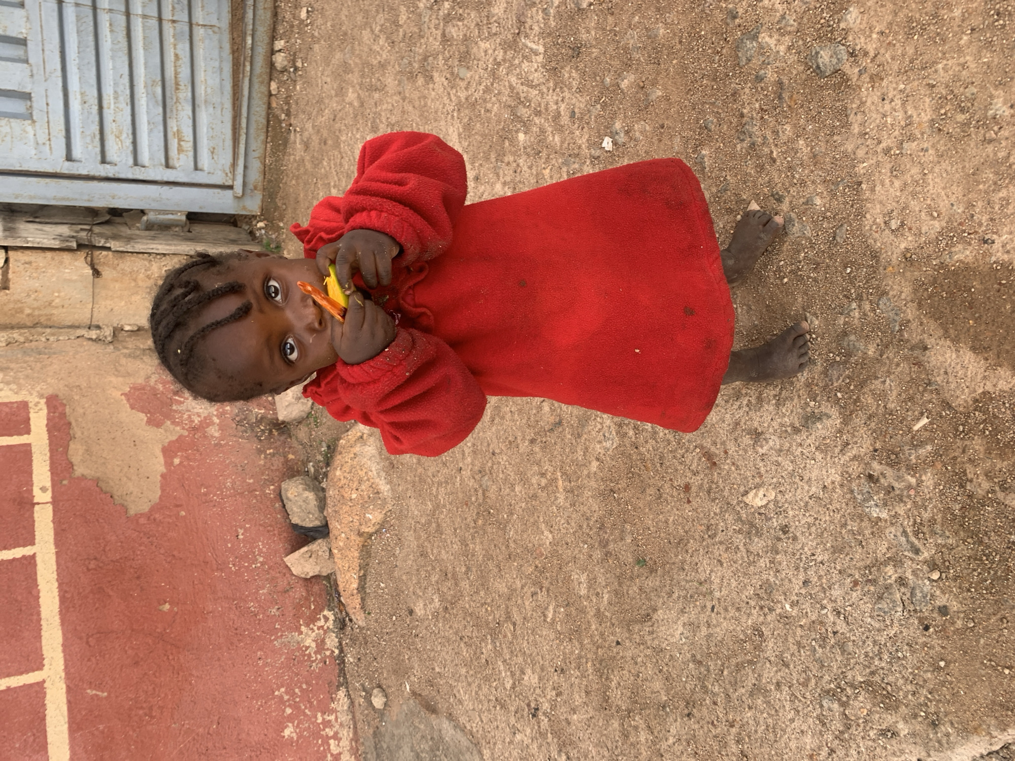 A young child with dark skin and braided hair is captured in a close-up, angled shot, looking directly at the camera with wide, engaging eyes. The child is wearing a bright red long-sleeved garment, possibly a fleece dress or top. They are holding a yellow and orange object, resembling a piece of fruit like a mango, near their mouth, as if eating or about to eat it.
The scene is outdoors, with the child standing barefoot on a varied ground surface. To the right, the ground is sandy and gravelly dirt, while to the left, it transitions to a reddish-brown concrete or paved area, marked with lighter patches and some visible stones. In the upper left background, a portion of a tall, grey metal gate with vertical slats is visible. The overall lighting suggests it is daytime, with dry conditions. The informal outdoor setting, clothing, and environment are consistent with daily life in a region such as Jos, Nigeria. There is no visible text in the image.