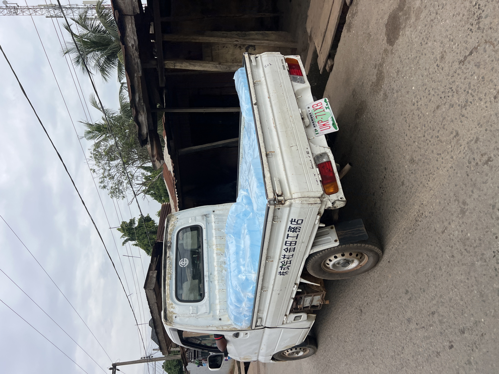 A white pickup truck is parked on an asphalt road in Nkpor, Nigeria. The truck's bed is covered with a bright blue tarp, beneath which is a load of what appears to be plastic-wrapped water bottles. The truck has a license plate that reads "UKP 71XB" and indicates the location as "ANAMBRA." The truck is positioned in front of a row of structures with corrugated metal roofs and rough-hewn wooden supports, suggesting a market or informal commercial area. Overhead, electrical wires crisscross the sky, and palm trees and other vegetation are visible in the background. The sky is overcast, and the overall lighting is subdued. No people are clearly visible in the image, though a hand wearing a red bracelet is slightly visible in the driver's side window.