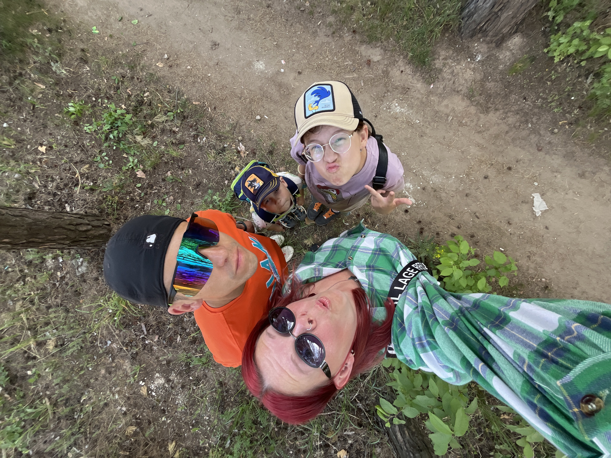 The image shows four people looking up at the camera. The person holding the camera is a woman with red hair, wearing sunglasses and a green plaid shirt. To her left is a person wearing an orange shirt and mirrored sunglasses. In front of these two are two children, both wearing hats and looking upwards. The older child is wearing glasses and making a face. The ground is visible with grass and a dirt path to the right of the image. The photograph appears to have been taken outdoors, possibly in a park or wooded area in the city of Chaikovsky, Russia.