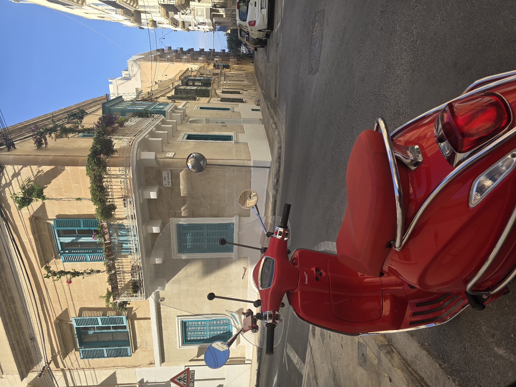 A bright red scooter is parked on the right side of a narrow asphalt street in Sliema, Malta. The scooter is positioned in the foreground, facing away from the camera and slightly to the left. Behind the scooter, the street curves to the right and uphill, lined with buildings. The buildings are constructed of light-colored stone and have traditional Maltese architectural features, including wooden shutters painted in shades of turquoise and green. Some buildings have balconies adorned with potted plants and flowers.
The sky above is clear and blue. Sunlight casts sharp shadows, indicating it is likely late morning or early afternoon. A traffic sign with a pedestrian crossing symbol and "25m" is visible on the left side of the street, near the corner. Further down the street, several cars are parked on the right. The overall scene depicts a typical streetscape in a Mediterranean city.