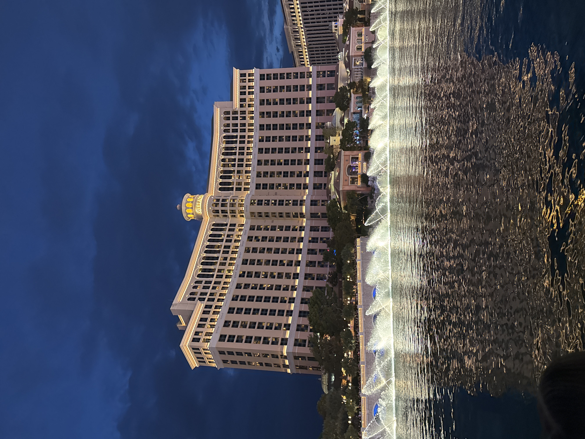 The image shows the Bellagio Hotel & Casino in Paradise, Nevada, United States of America, at dusk or night. The hotel is a large, light-colored building with multiple floors and arched windows. It is illuminated by warm lighting, highlighting architectural details and the "Bellagio" sign on its central tower. In front of the hotel is a large body of water with a fountain display. The fountains are spraying water illuminated with white and blue lights, creating a dynamic visual effect. The water reflects the hotel's lights and the dark, cloudy sky. The environment appears to be a public area, likely a promenade or viewing area, with some trees and smaller structures visible behind the fountains and in front of the hotel. The overall scene depicts a well-known tourist attraction, showcasing a blend of impressive architecture and water features.