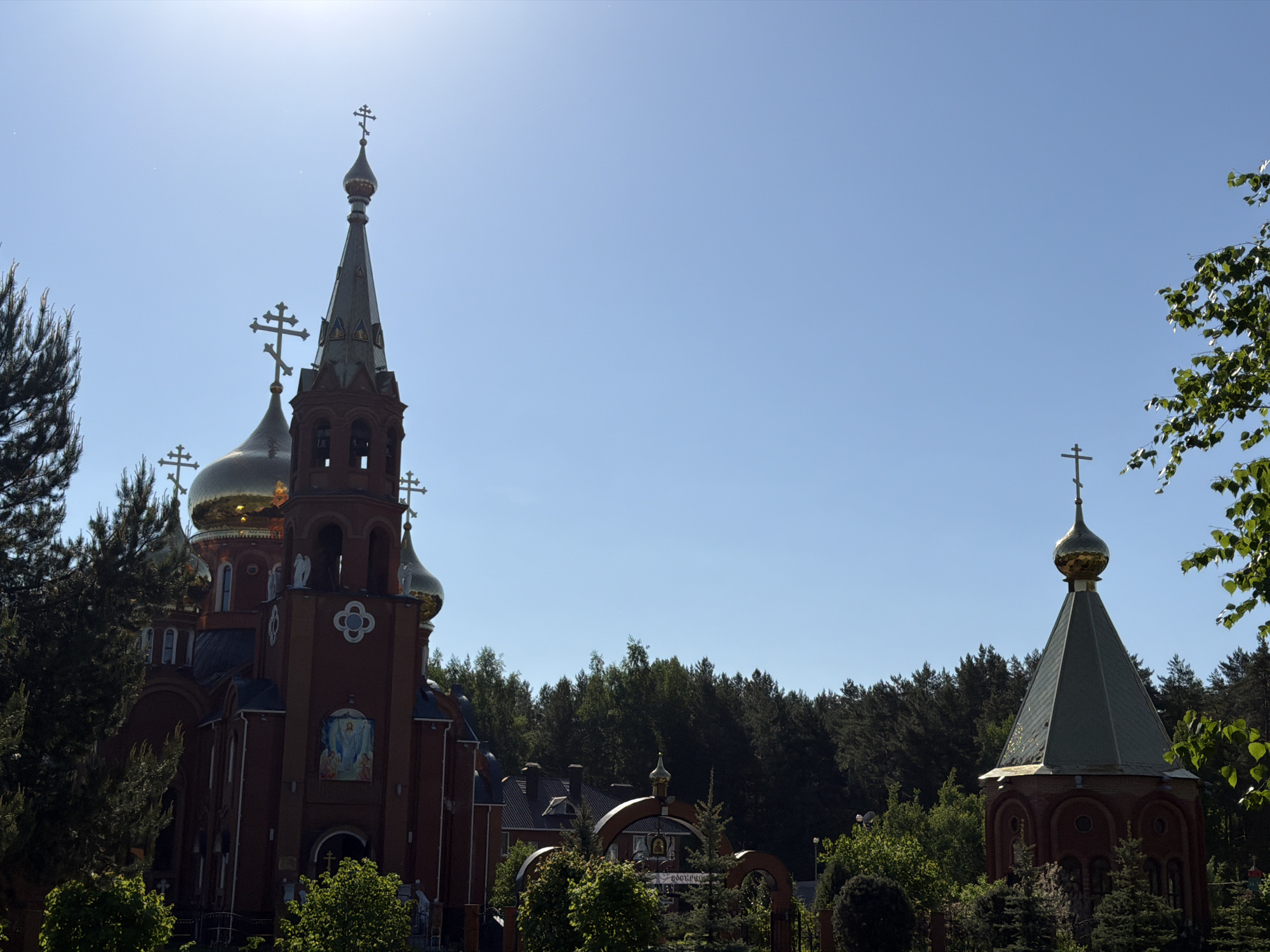 The image depicts a Russian Orthodox church complex under a clear, blue sky in Chaikovsky, Russia. The main church features a red brick facade with golden onion domes topped with Orthodox crosses. Adjacent to the main church is a taller, thinner bell tower, also made of red brick and topped with a spire and a cross. To the right of the main church, partially obscured by trees, is a smaller chapel with a conical roof and a golden dome surmounted by a cross. The church complex is surrounded by trees and greenery, with a dark green forest visible in the background. The sun is shining brightly, causing some glare in the sky. A gate is also visible in between the structures.